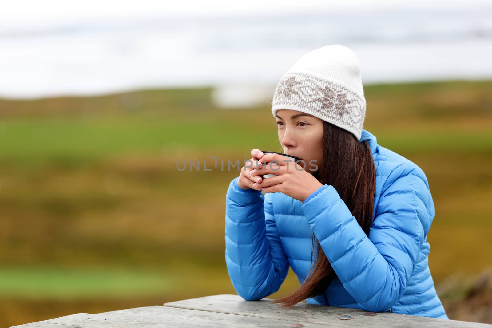 Woman in outdoors drinking coffee from thermos cup sitting outside wearing warm down jacket and knit hat. Pretty young mixed race Asian Chinese Caucasian woman living active lifestyle.