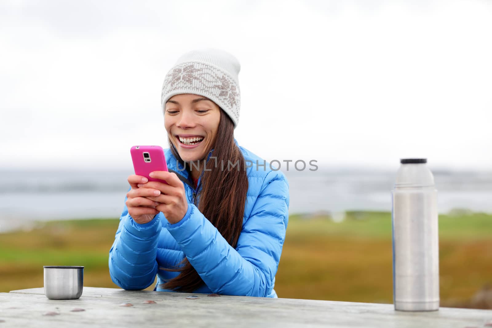 Woman outdoors using smartphone app on smart phone drinking coffee from thermos cup sitting outside wearing warm down jacket. Pretty young mixed race Asian Chinese Caucasian woman in active lifestyle.