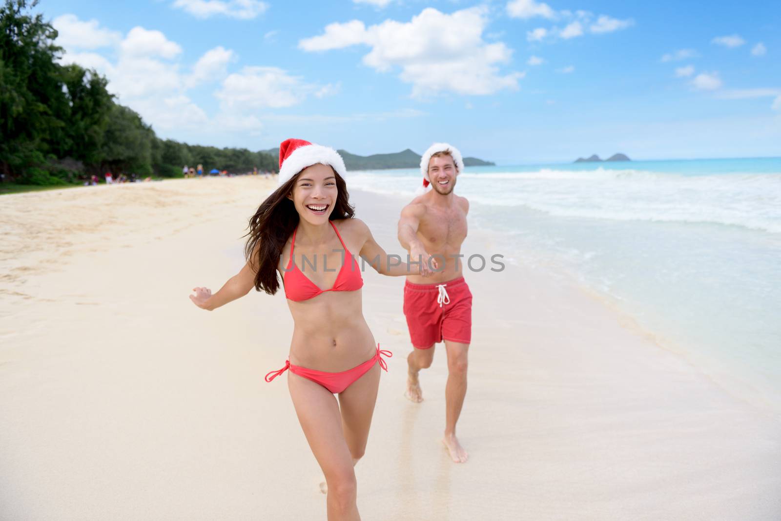 Christmas couple happy relaxing on white sand beach running on sand in bikini and swimsuit. Asian woman and man holding hands running playing during travel holidays.