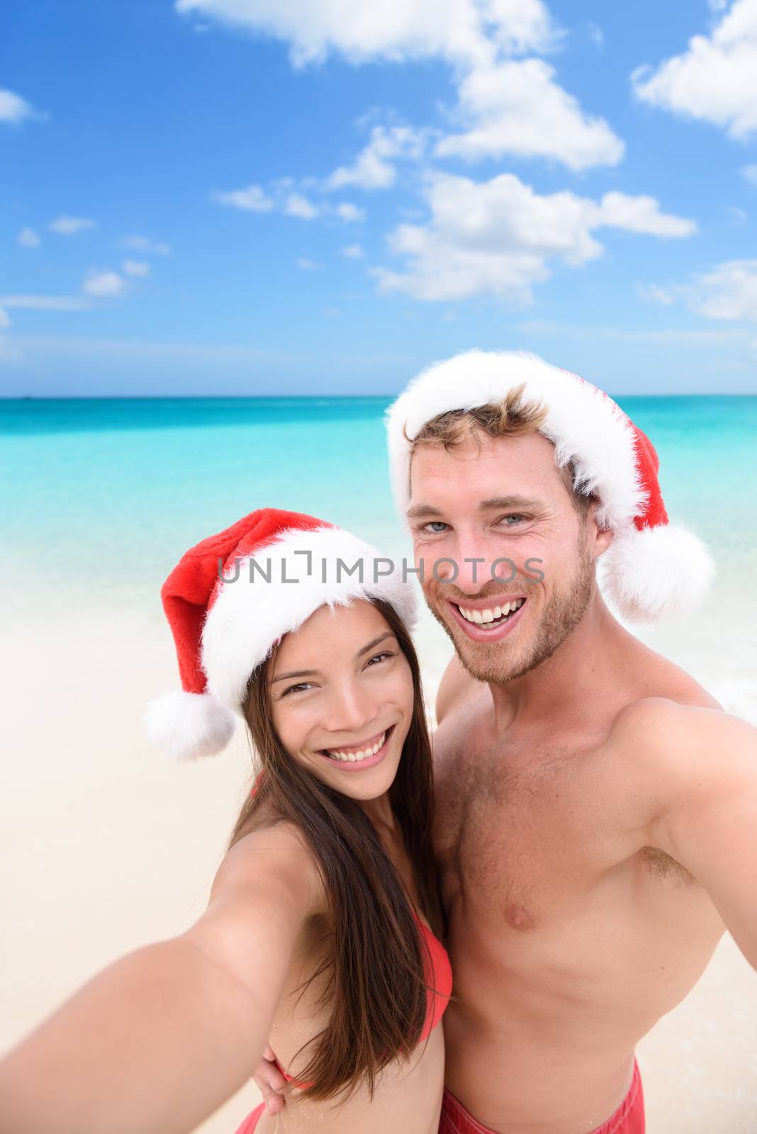 Happy couple on Christmas travel holidays taking selfie picture with smartphone wearing santa hat during their winter vacation. Young adult friends in swimsuit and bikini in front of the ocean.