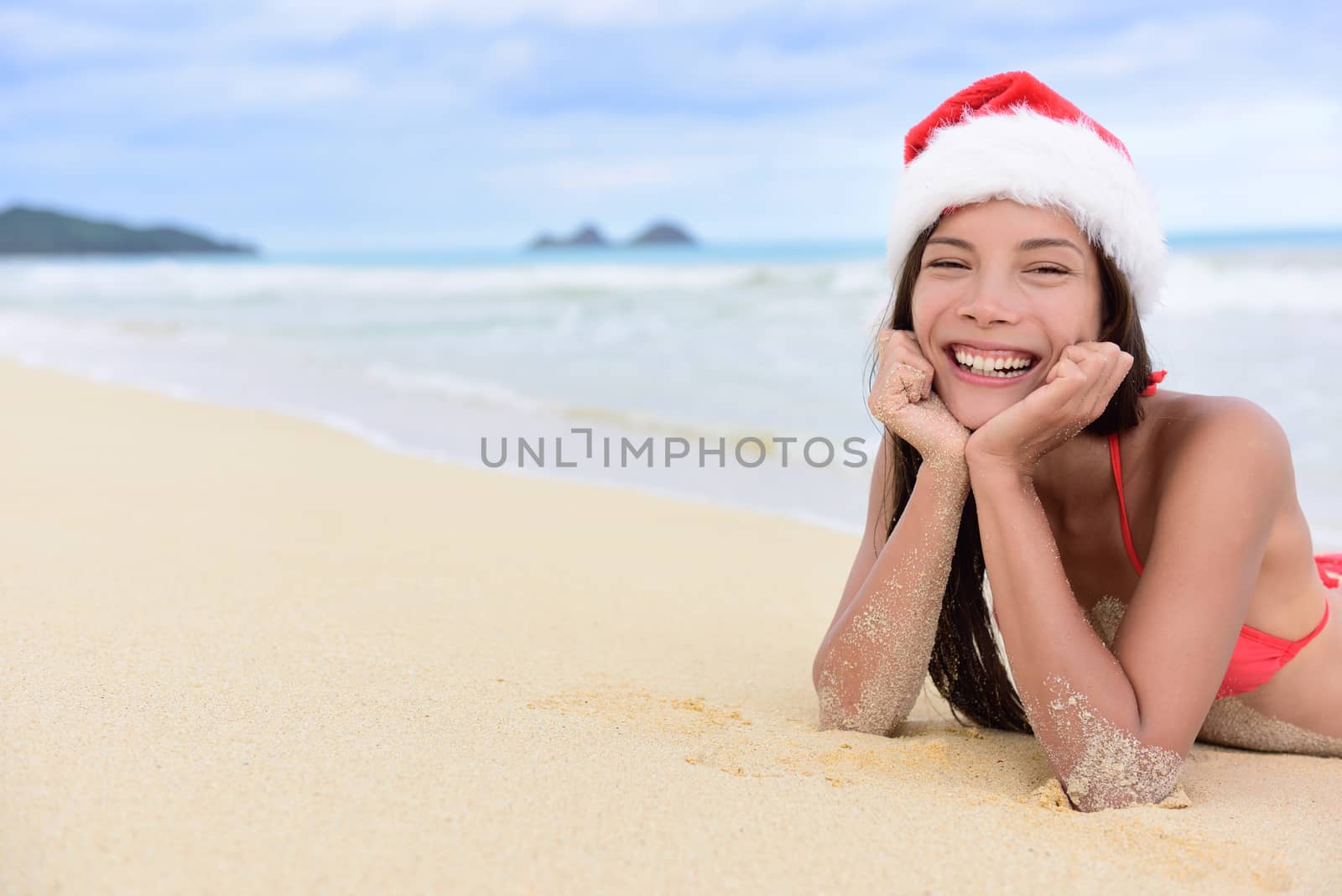 Christmas beach vacation - cute mixed race Asian Chinese girl portrait in Santa hat lying down relaxing posing on white sand in a tropical destination during winter holidays.