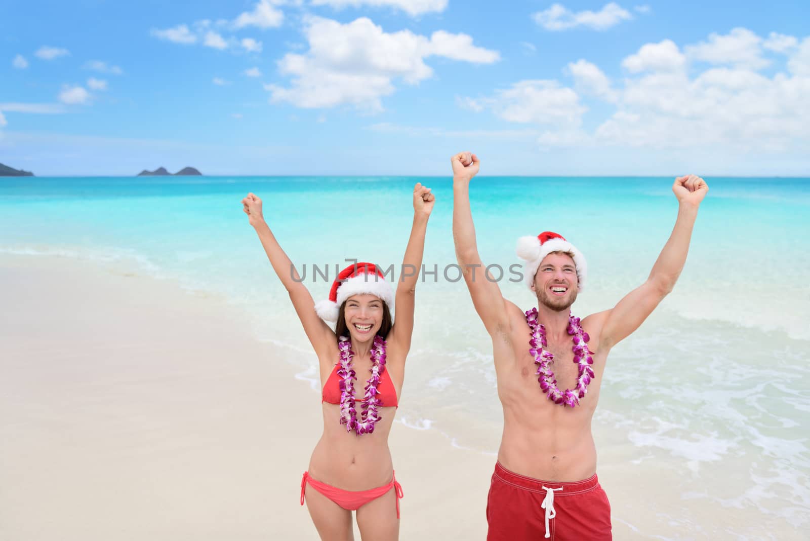 Happy Christmas holiday - multiracial joyful couple cheering arms up on Hawaii beach for their winter vacation during new year.