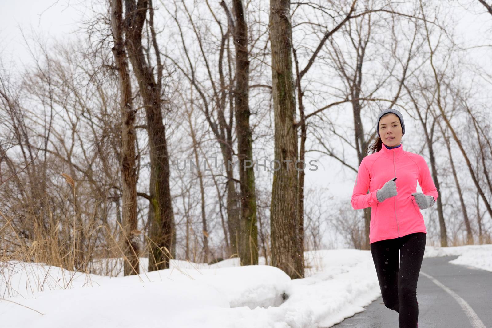 Winter cardio exercise - woman jogging doing her workout outside. Young adult running in outdoor park with snowy forest background wearing cold weather gear.