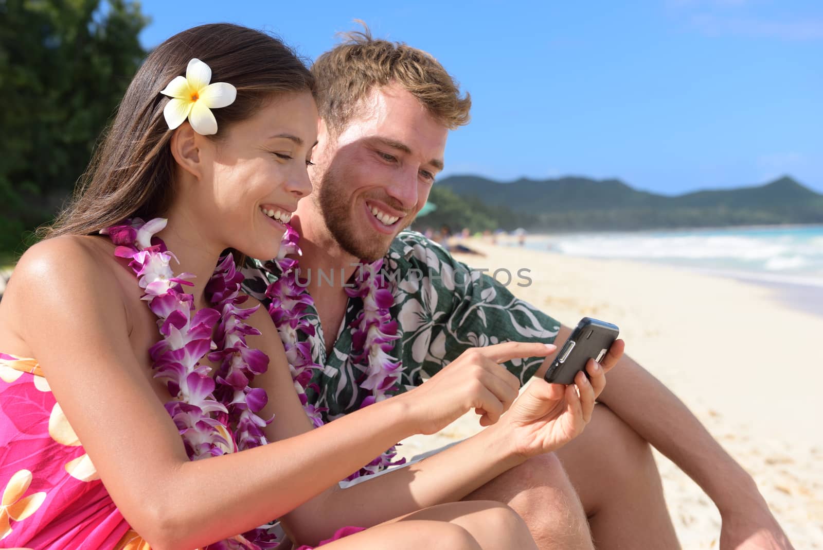 Couple on beach using smart phone app sharing photos on social media on Hawaii with smart phone. Young woman and man in love on beach vacations in Hawaiian clothing flower lei.