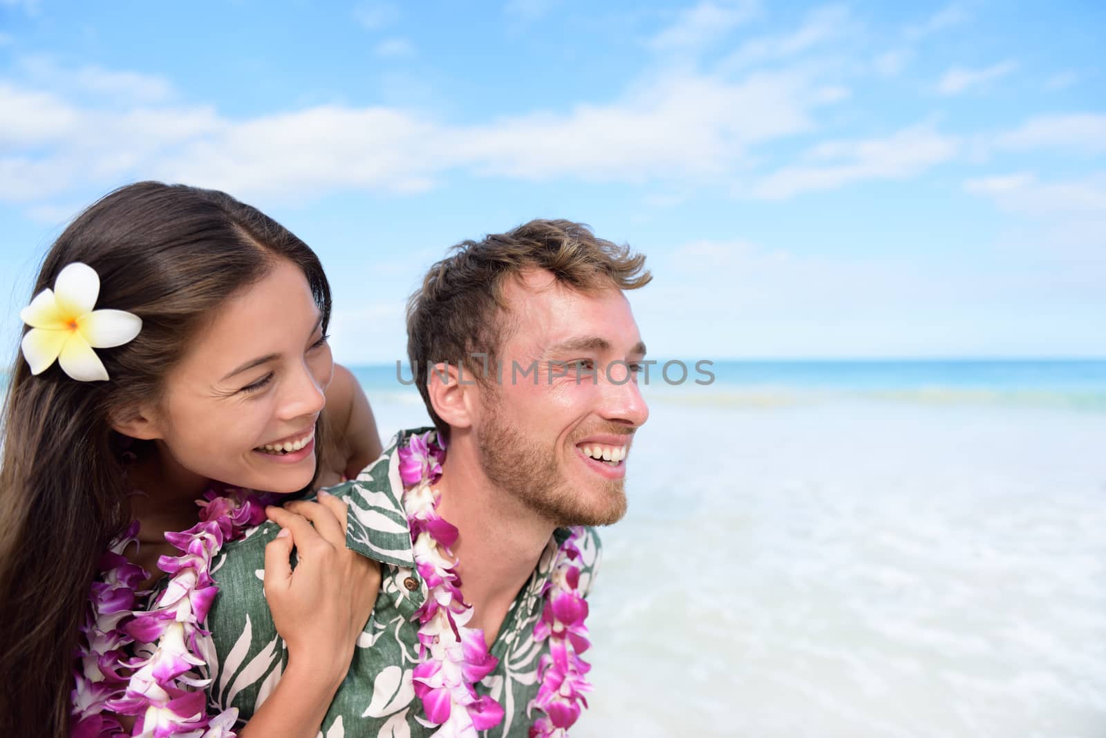 Beach couple having fun piggybacking and laughing on Hawaii travel holiday. Beautiful Asian mixed race woman piggybacking on Caucasian boyfriend wearing traditional Hawaiian lei on Oahu, Hawaii, USA.