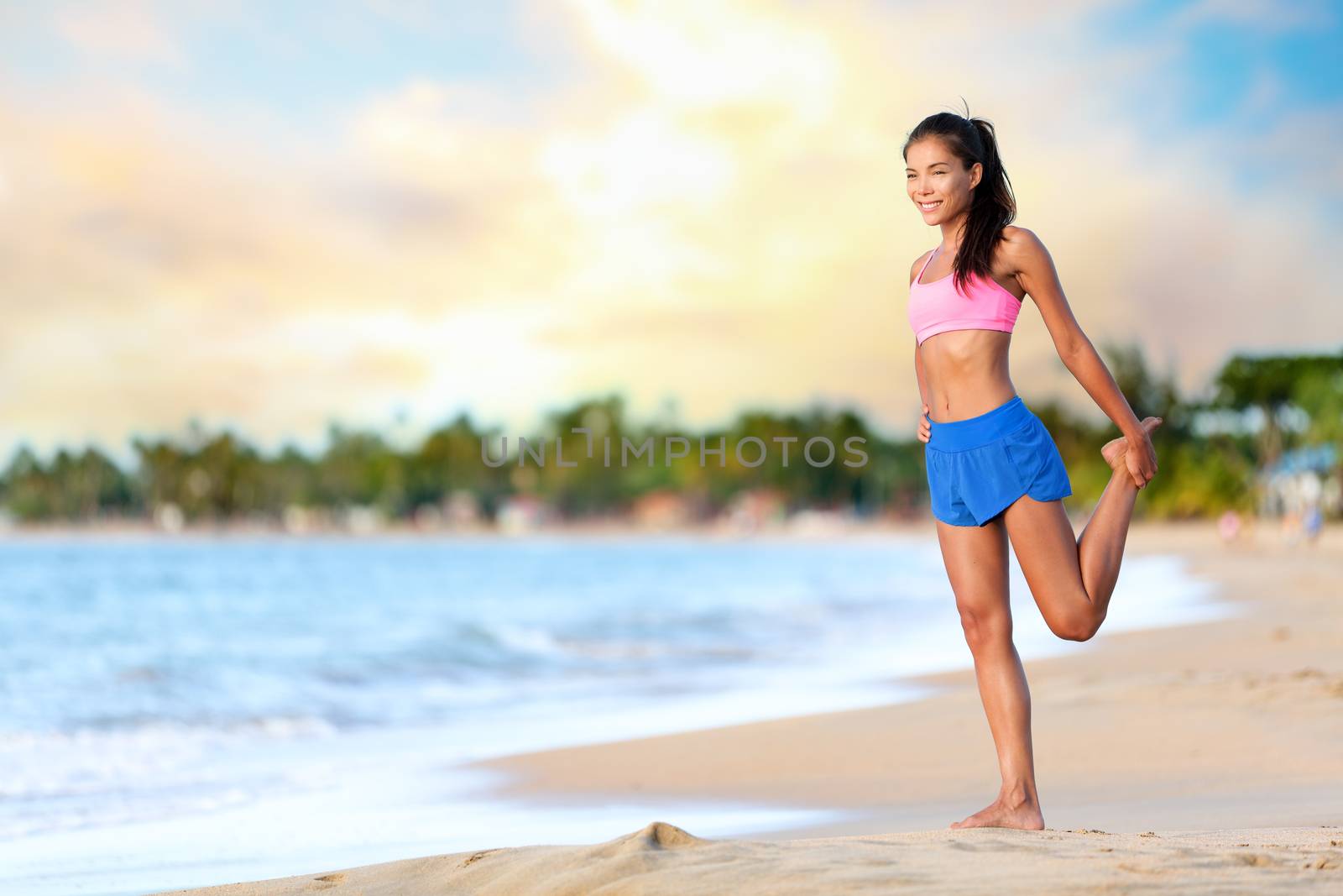 Happy young woman doing stretching exercise on beach. Smiling female is wearing sports bra and shorts. Full length of sporty woman looking away while exercising on sea shore against cloudy sky.