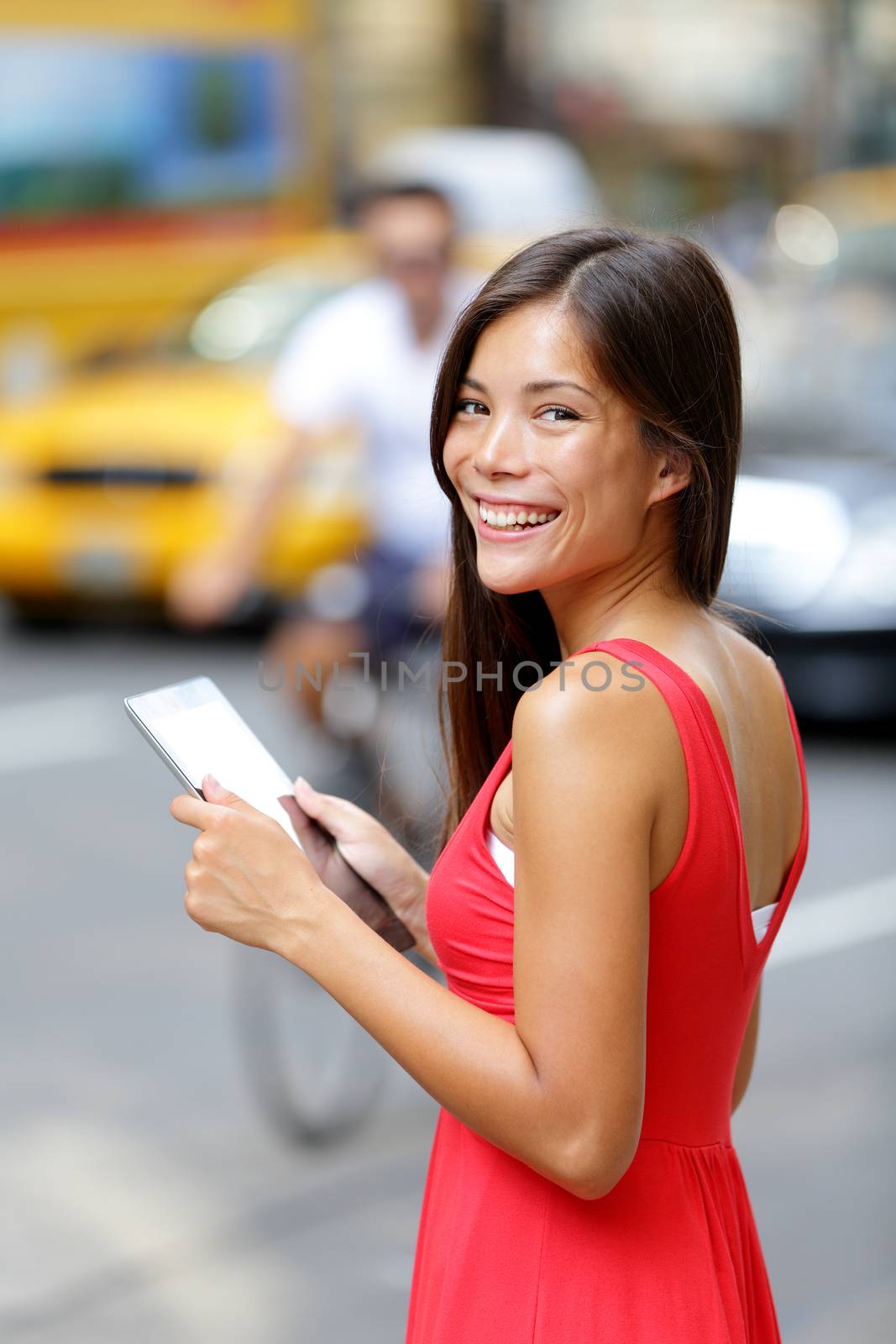 Happy woman holding digital tablet while standing on city street. Side view of young female is wearing red dress. Smiling beautiful woman is with tablet PC on street in city.