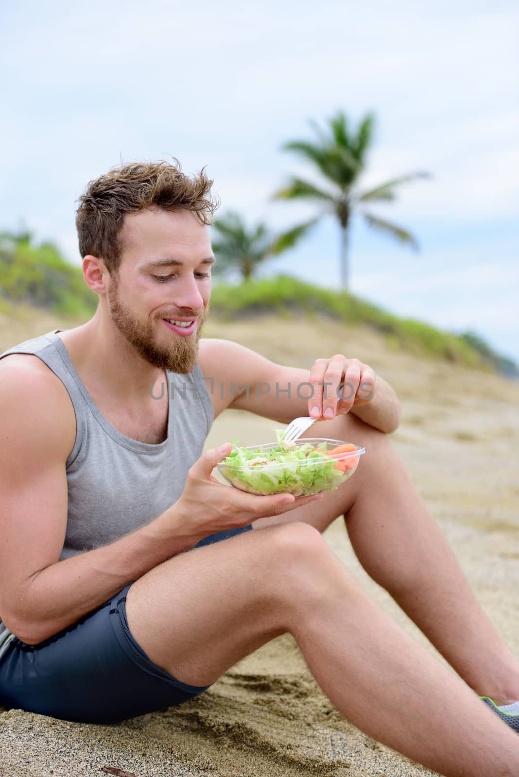 Healthy fit man on vegan diet eating organic food on beach. Muscular young fitness guy after exercise eating take-out food of prepared fresh salad and veggies. by Maridav