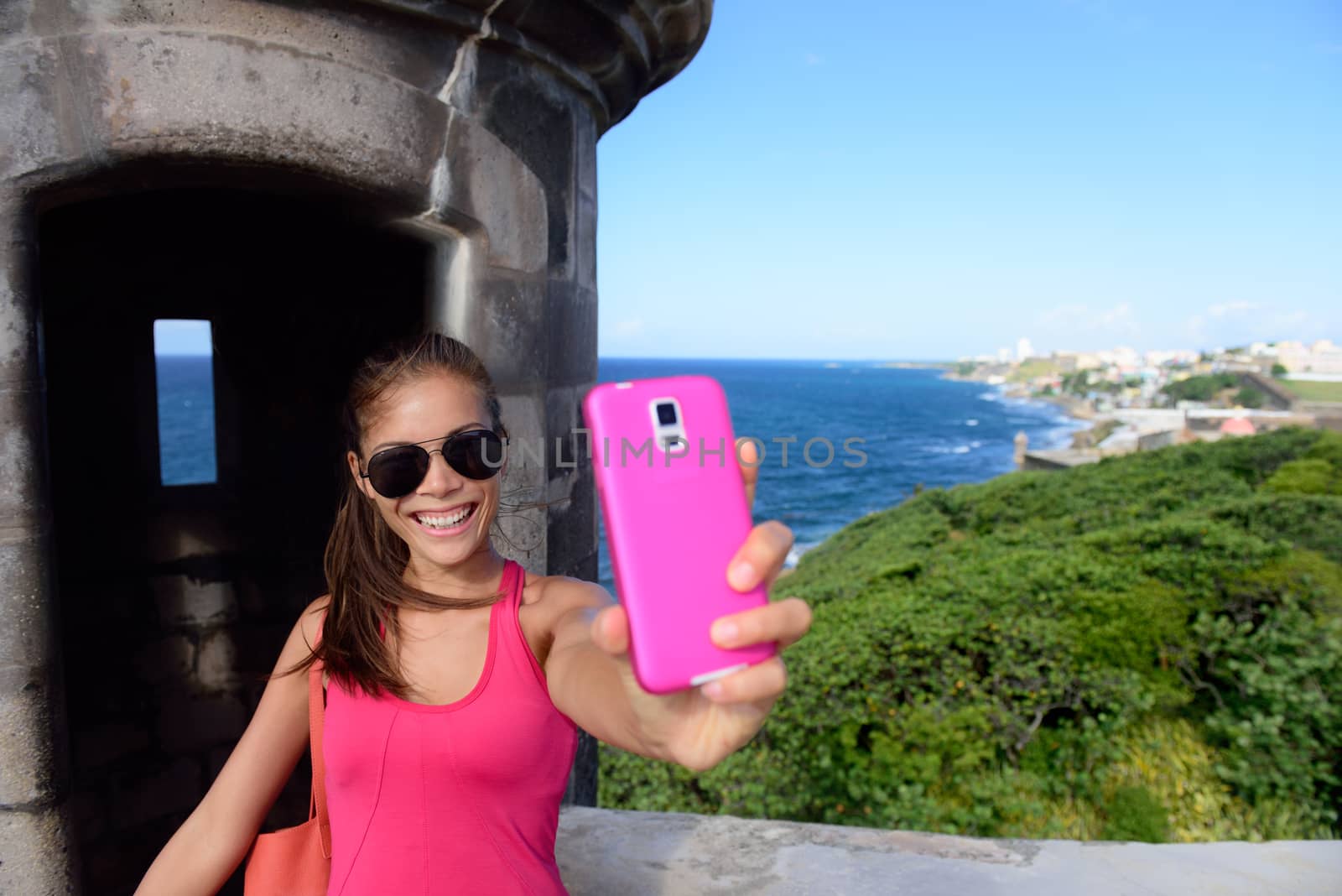 Tourist taking fun selfie at famous landmark. Travel woman holding a pink smartphone visiting Old San Juan's Castillo San Felipe Del Morro, the main attraction of the city of San Juan, Puerto Rico.