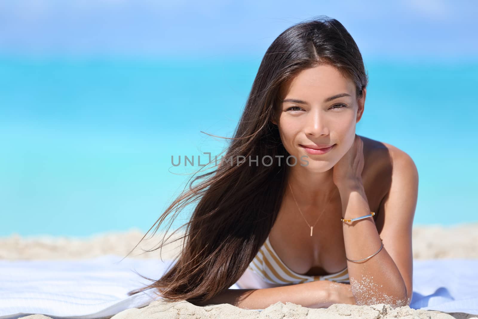 Asian woman wearing bikini and jewelry - bracelet and necklace - relaxing on beach. Portrait of Chinese Caucasian multiracial fashion model lying down on white sand on Caribbean vacation travel.