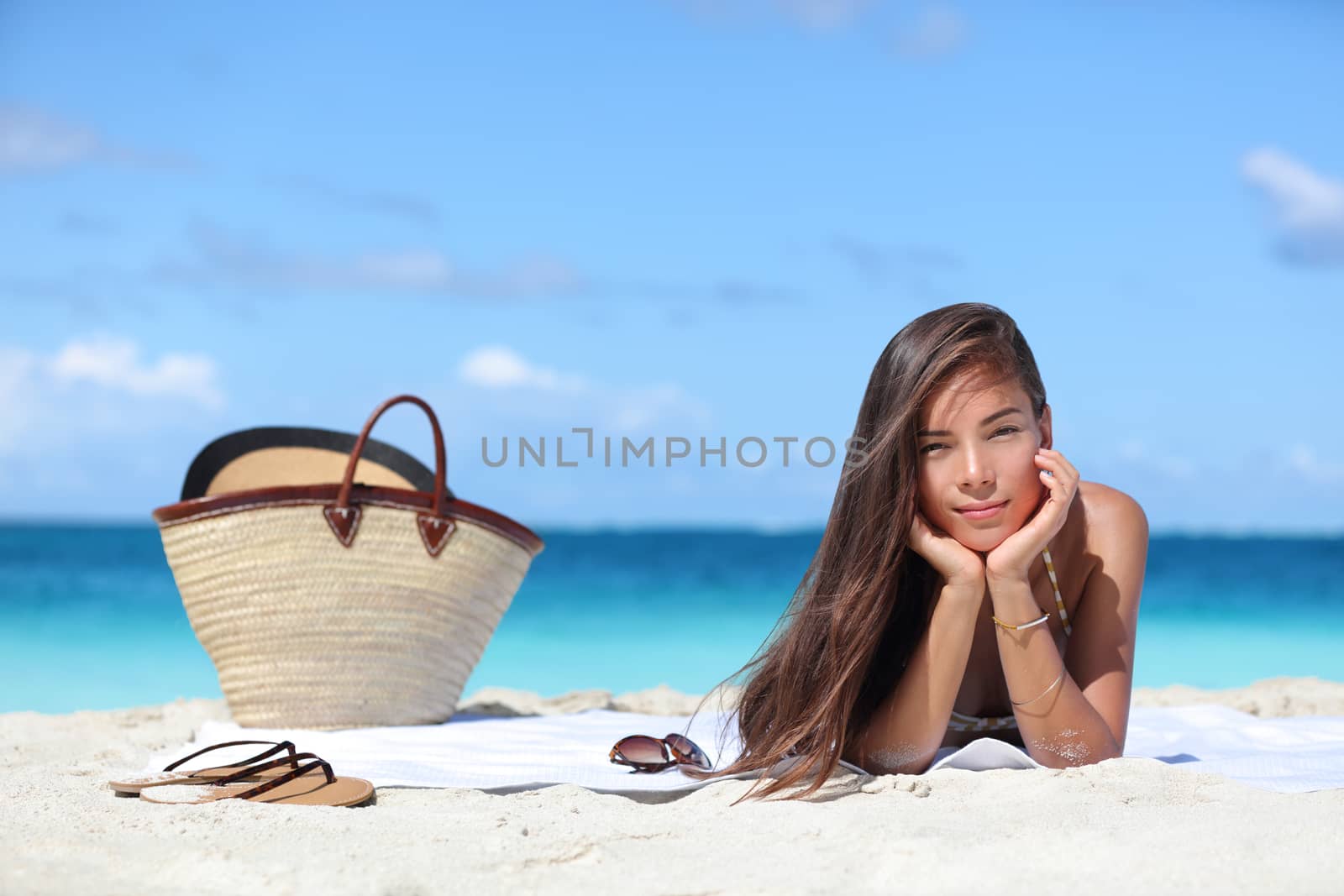 Woman relaxing on beach vacation summer holidays. Beautiful Asian girl lying down on towel with straw hat and flips flops on white sand tanning. Sun hair and skin care concept.