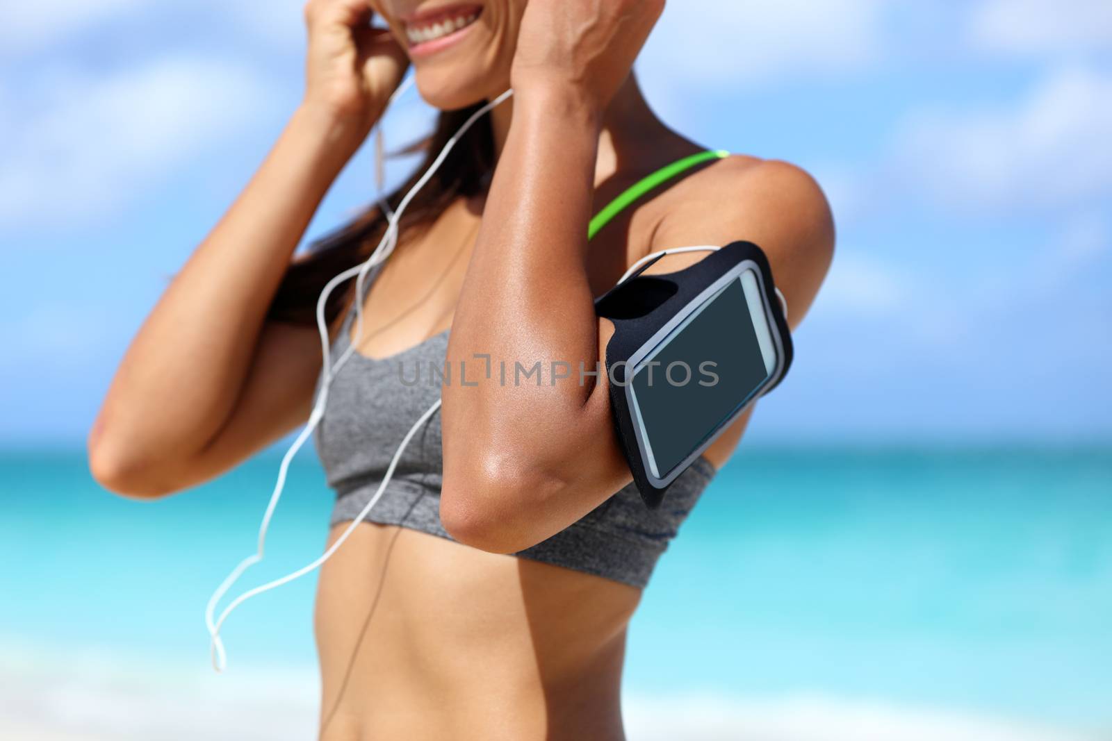 Fitness phone armband runner woman putting earphones. Closeup of sports smartphone case holder touchscreen strap on female arm of person wearing headset for running exercise cardio workout on beach.