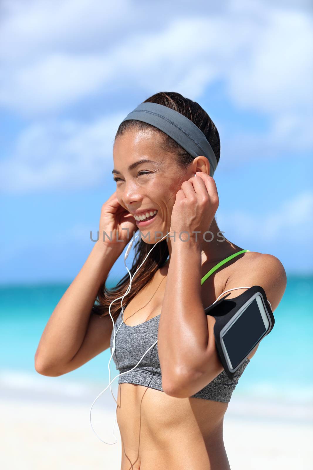 Runner girl wearing earphones and running armband phone arm strap getting ready for run workout. Gesture of putting on headset to listen to music while exercising on sunny beach summer vacation.