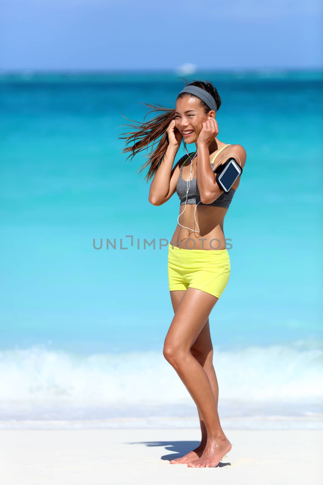 Running woman wearing earphones and phone arm strap with touchscreen listening to music for workout motivation. Female athlete standing full body on beach putting on in-ear headphones for exercise.