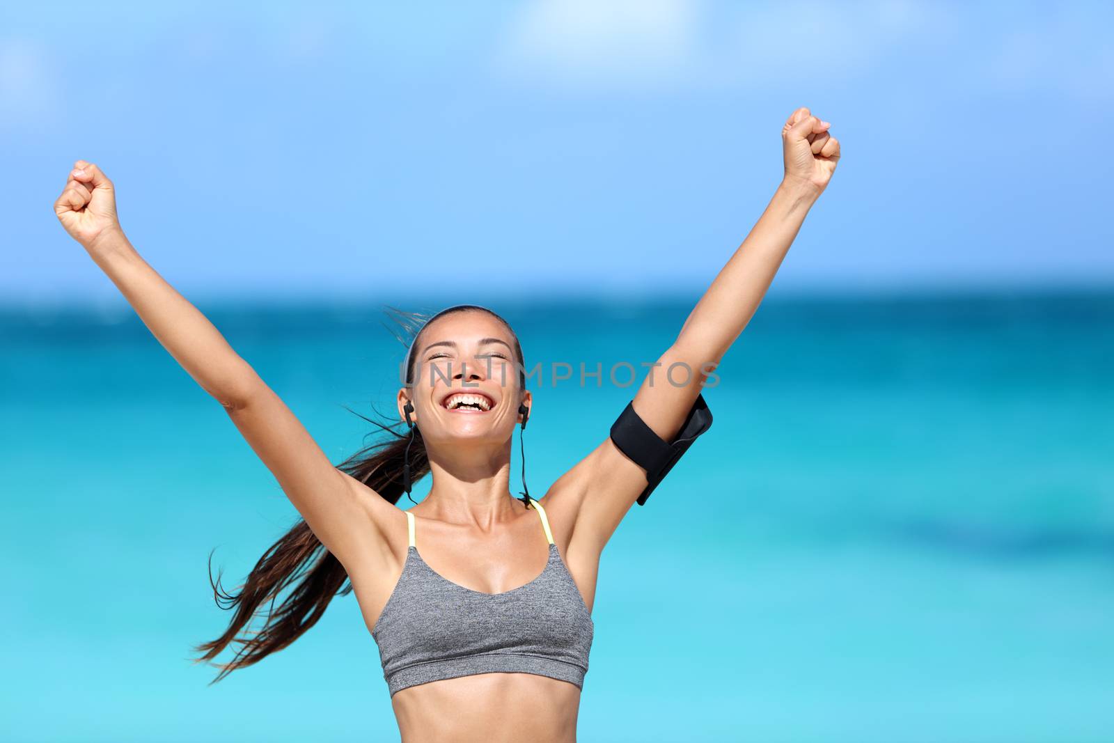 Happy running woman winning - fitness goal concept. Young Asian female runner smiling of happiness cheering with arms up wearing a sports bra, earphones and phone armband cheering of success.