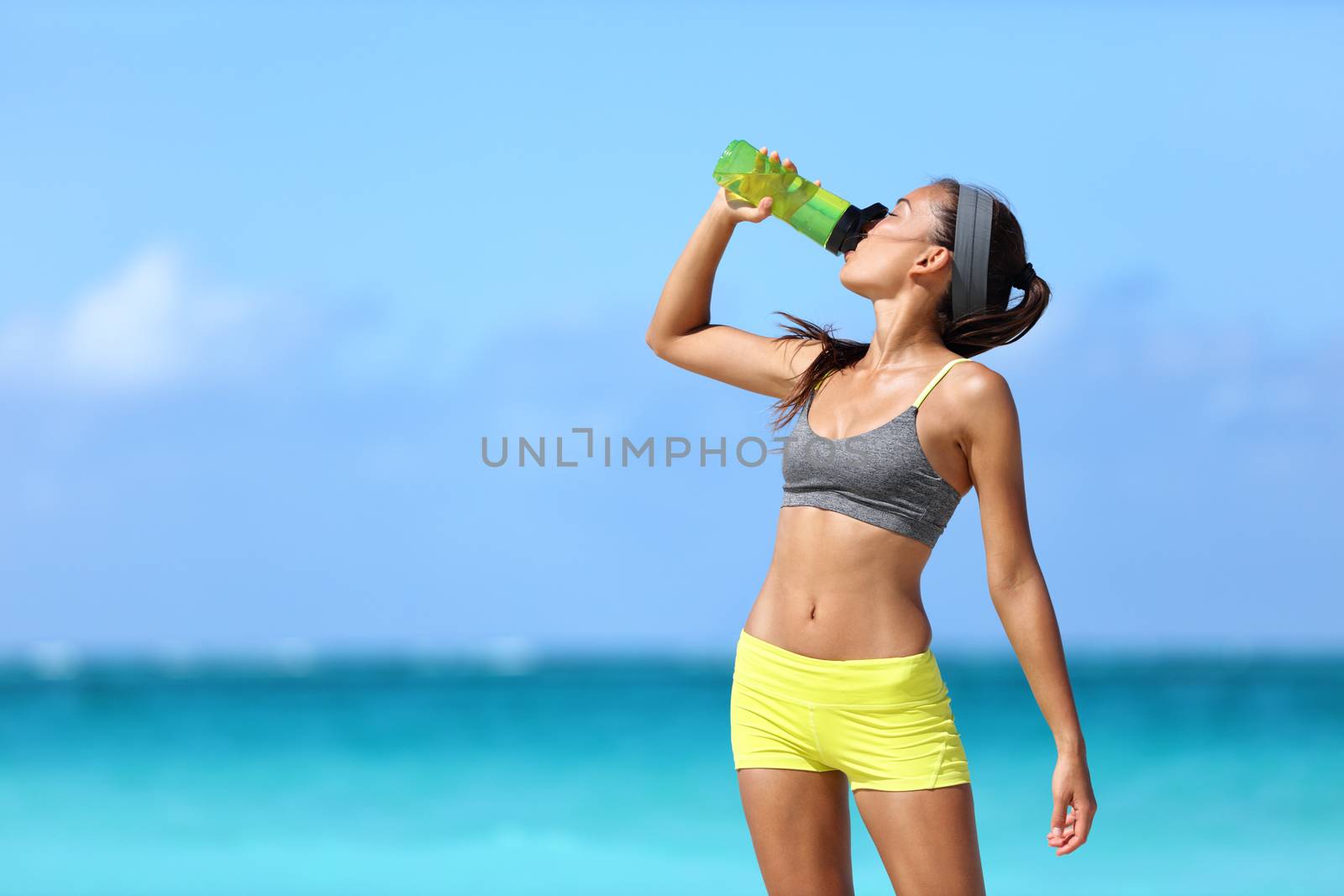 Fitness runner woman drinking water or energy drink of a sport bottle. Athlete girl taking a break during run to hydrate during hot summer exercise on beach. Healthy active lifestyle.