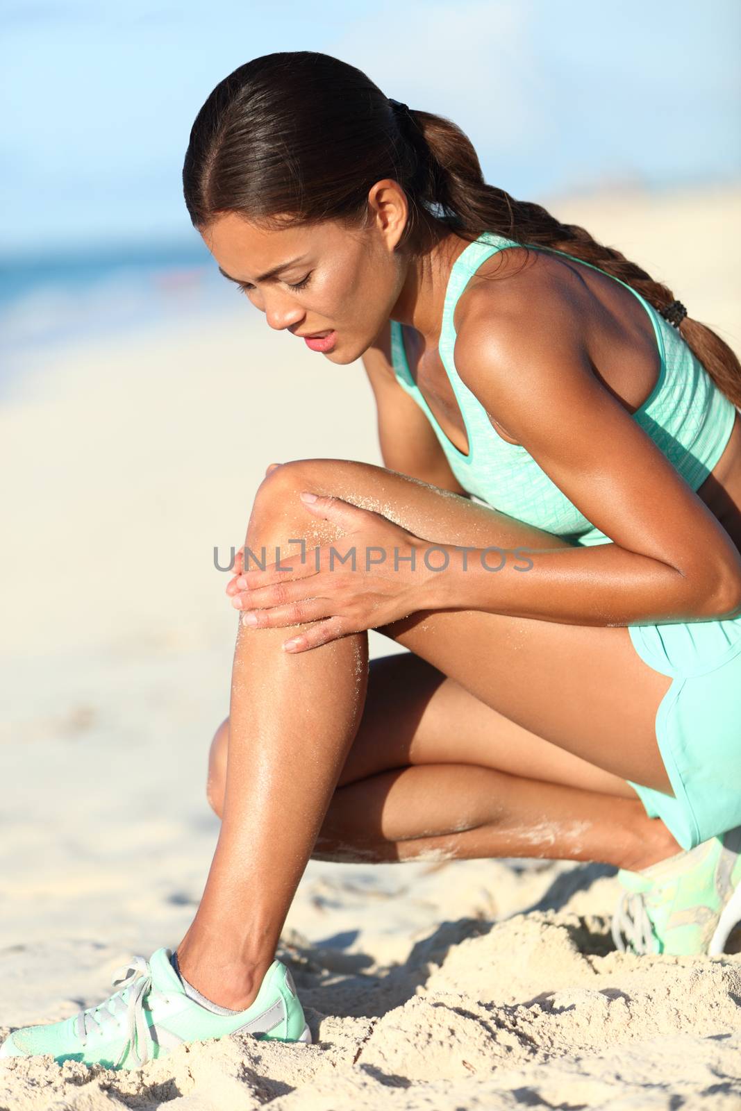 Runner injury - running woman upset with hurting knee pain. Asian female athlete with painful legs holding her leg in ache because of sport problem. Sporty girl jogging on beach.