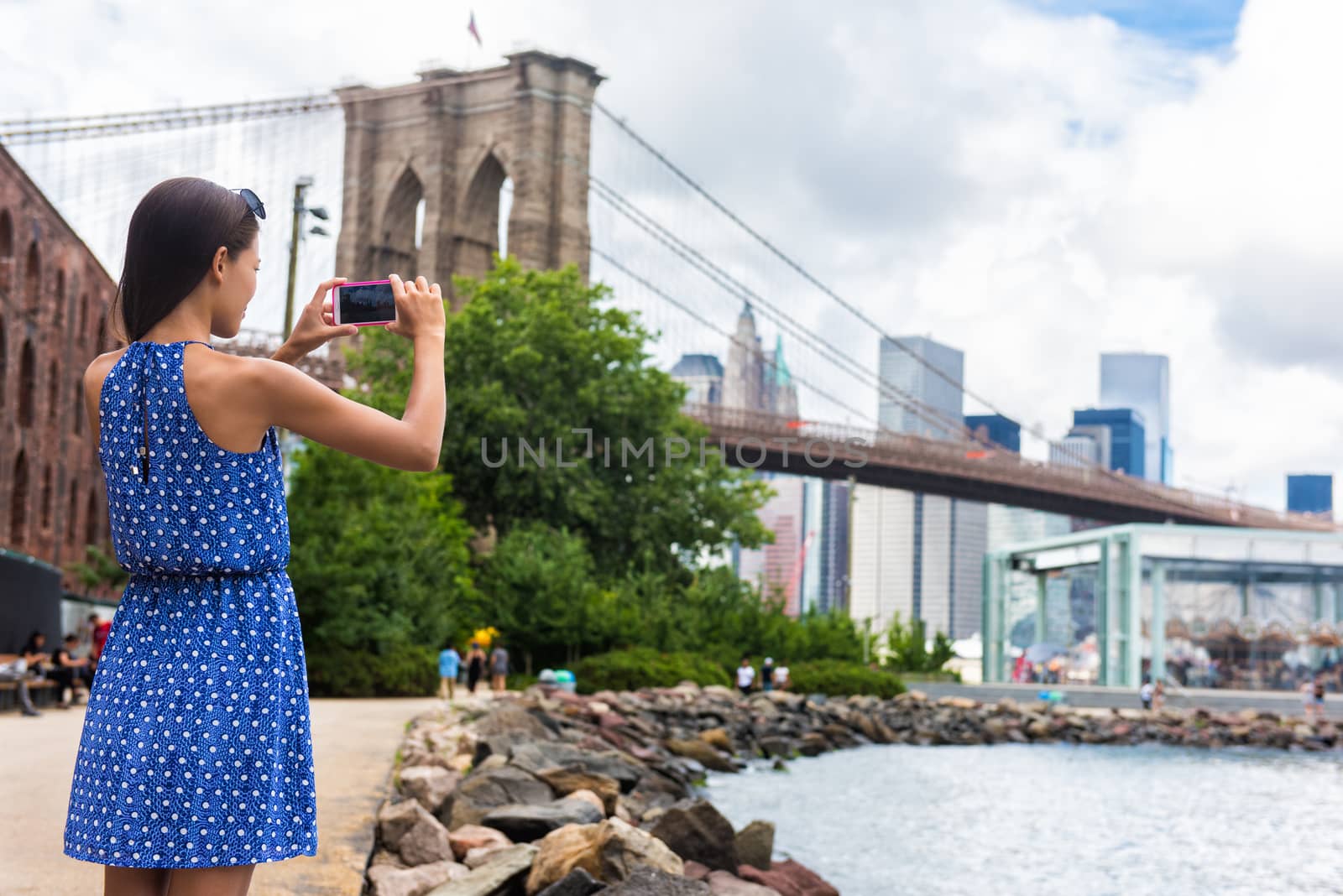 Tourist taking travel picture with phone of Brooklyn bridge and New York City skyline during summer holidays. Unrecognizable female young adult enjoying USA vacations in blue dress.