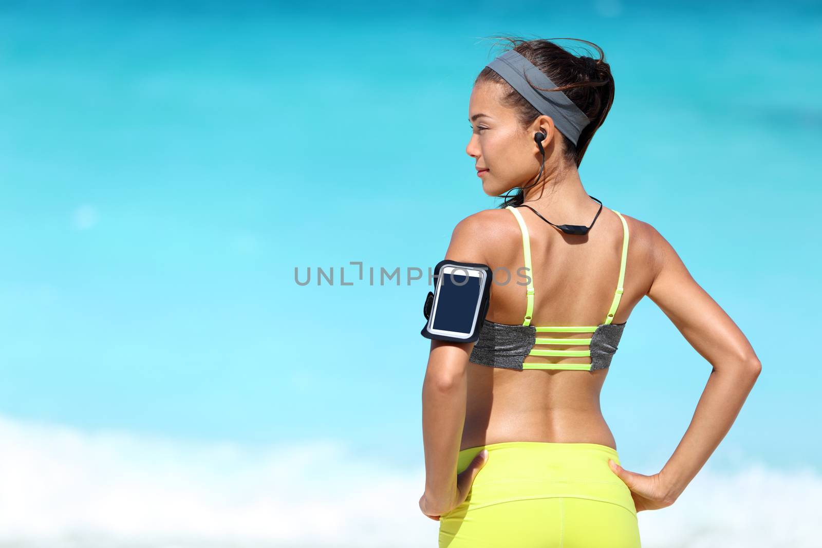 Fitness runner woman with fit back wearing phone armband and wireless headphones on ocean background. Multiracial exercise girl jogging on summer beach with sports smartphone touchscreen for app.