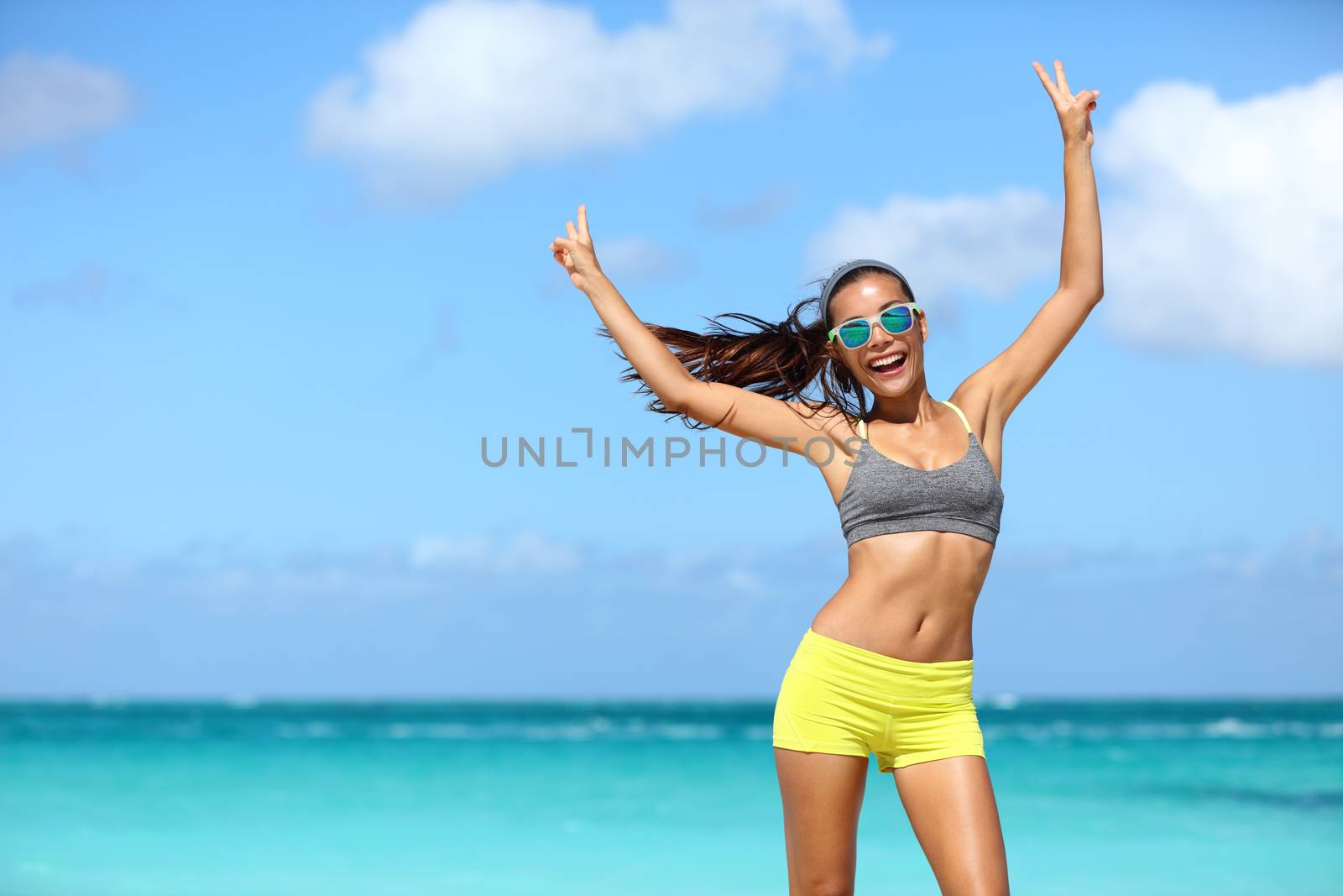 Happy cheerful fitness woman in sunglasses winning arms up doing v hand signs. Fit girl with slim body on sunny beach with raised arm showing peace sign in happiness of weight loss after workout.