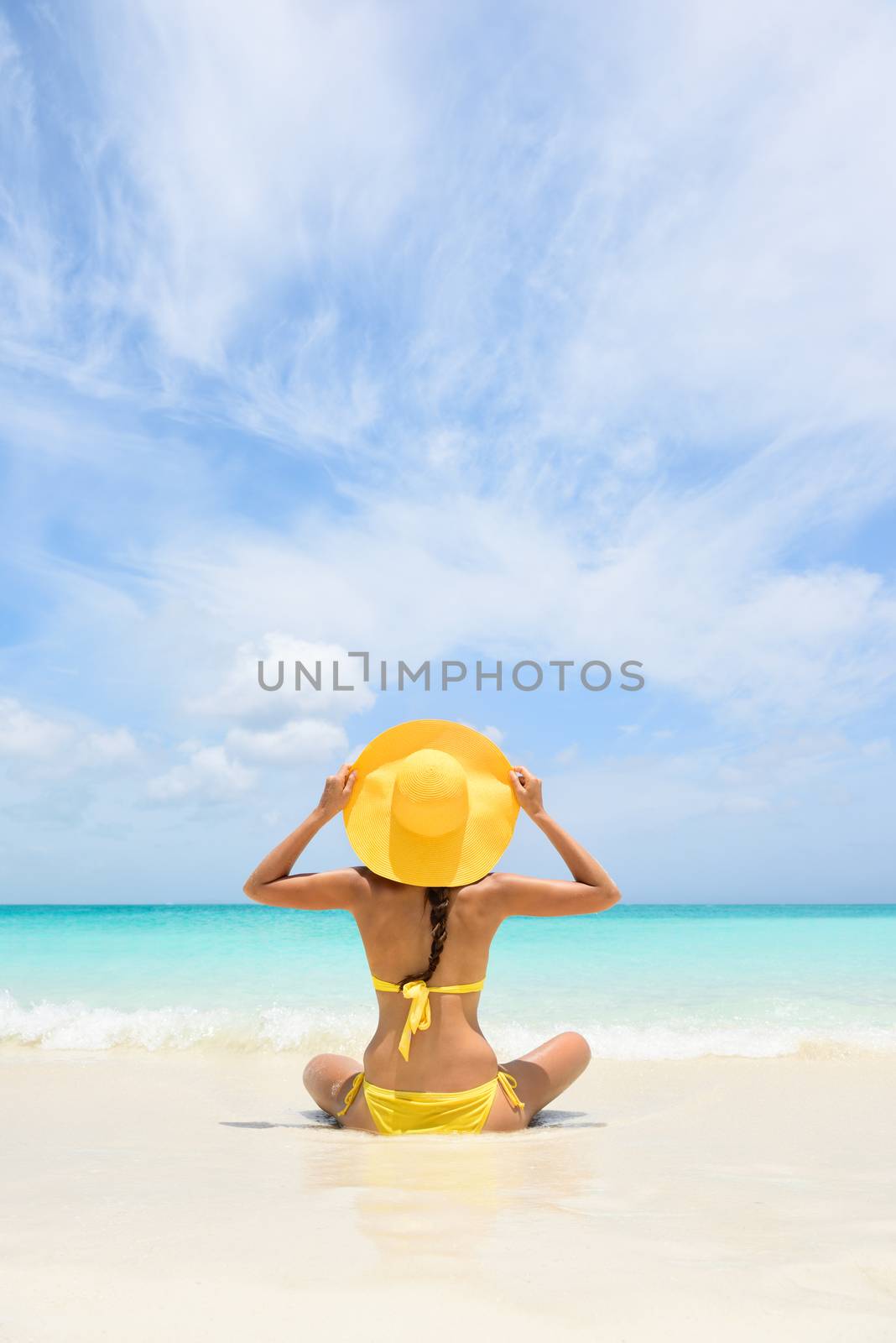 Beach Travel Vacation Holidays Woman Relaxing. Woman sitting on sea shore during summer. Rear view of female wearing yellow sunhat and bikini. Carefree tourist is enjoying vacation at beach.