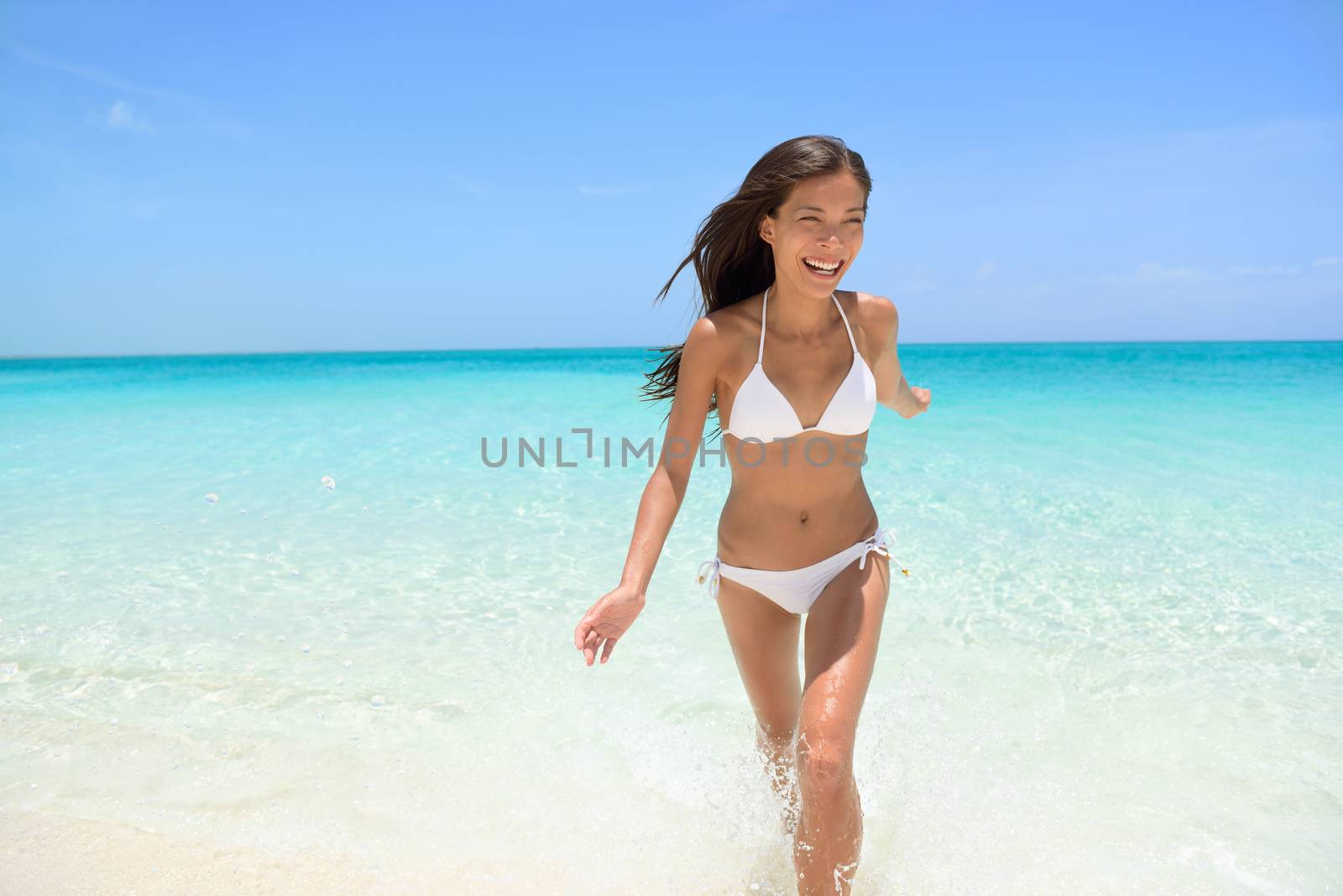 Cheerful young woman running on beach having fun laughing during summer holidays travel. Exhilarated female is in white bikini smiling. Beautiful tourist is enjoying vacation on beach.