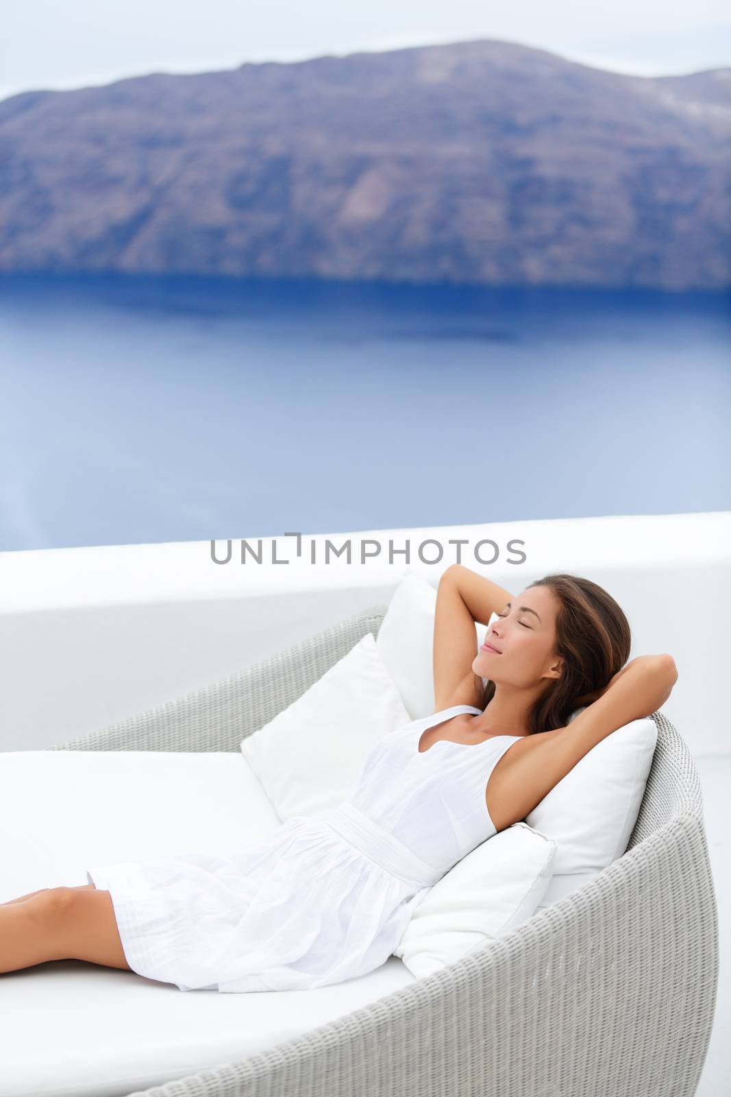 Beautiful young woman relaxing on couch at seaside resort terrace. Attractive female is wearing white sundress. Female with hands behind head is relaxing on lounge chair against sea and mountain.