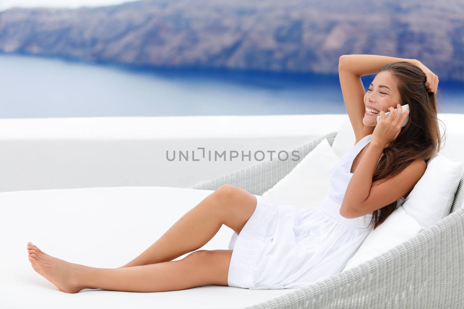 Happy young woman using smartphone while relaxing on couch. Smiling female talking on smart phone is at seaside resort terrace. Female tourist is wearing white sundress.
