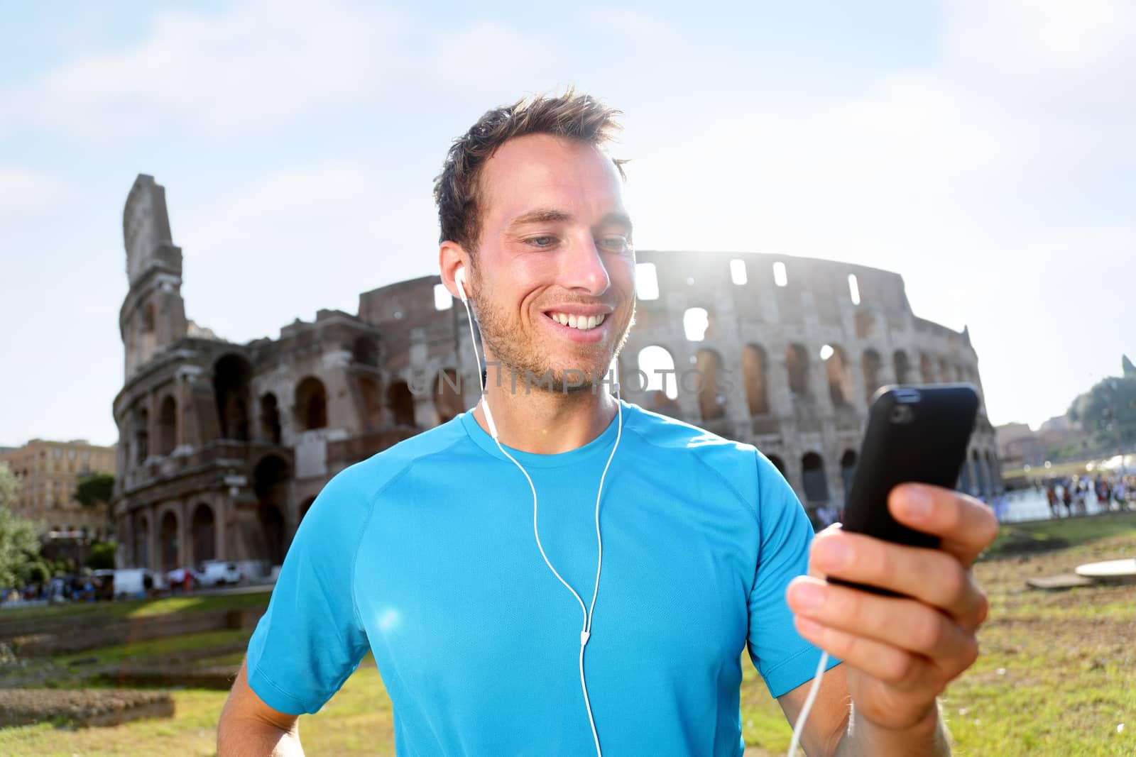 Happy male jogger listening music on smartphone against Colosseum. Smiling man is in sportswear. Handsome fit runner is enjoying music before workout on sunny day.