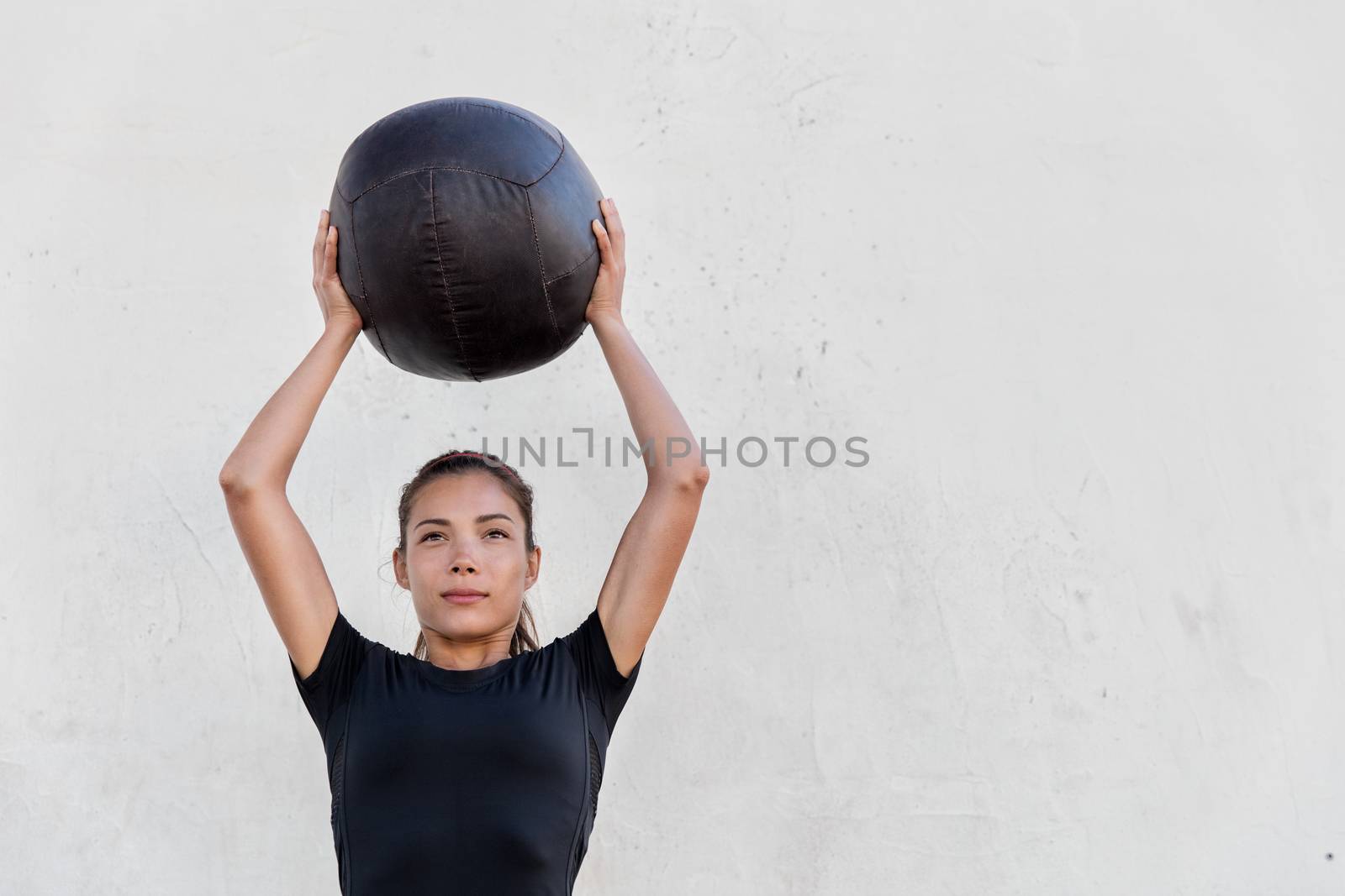 Fitness crossfit girl holding medicine ball above head for shoulder press workout in outdoor crossfit gym. Young Asian athlete girl doing upper body exercise working out with heavy weighted balls.
