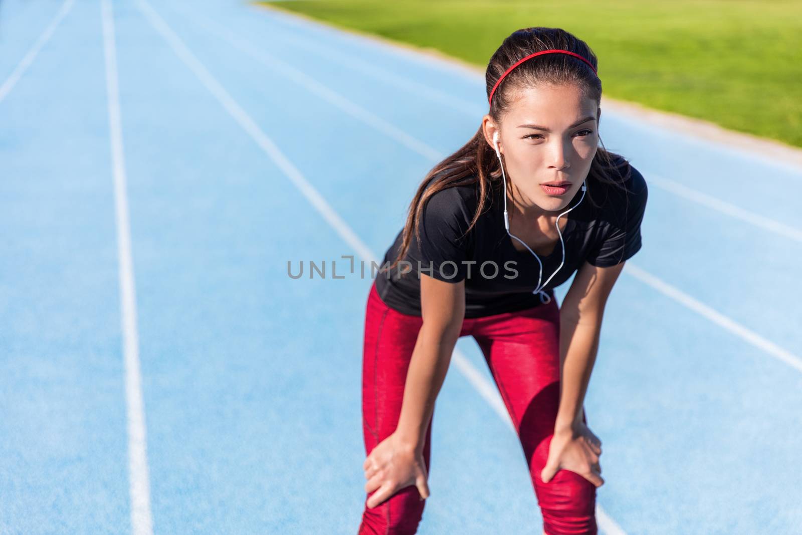 Runner resting on outdoor running track tired taking a break getting determination and motivation ready for the challenge. Athlete woman listening to music with earphones for focus.