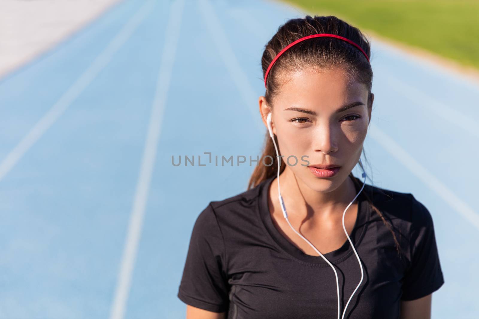 Runner woman listening to music with earphones for running motivation getting ready to race. Asian female athlete training focus, determination and cardio on athletic tracks at stadium.