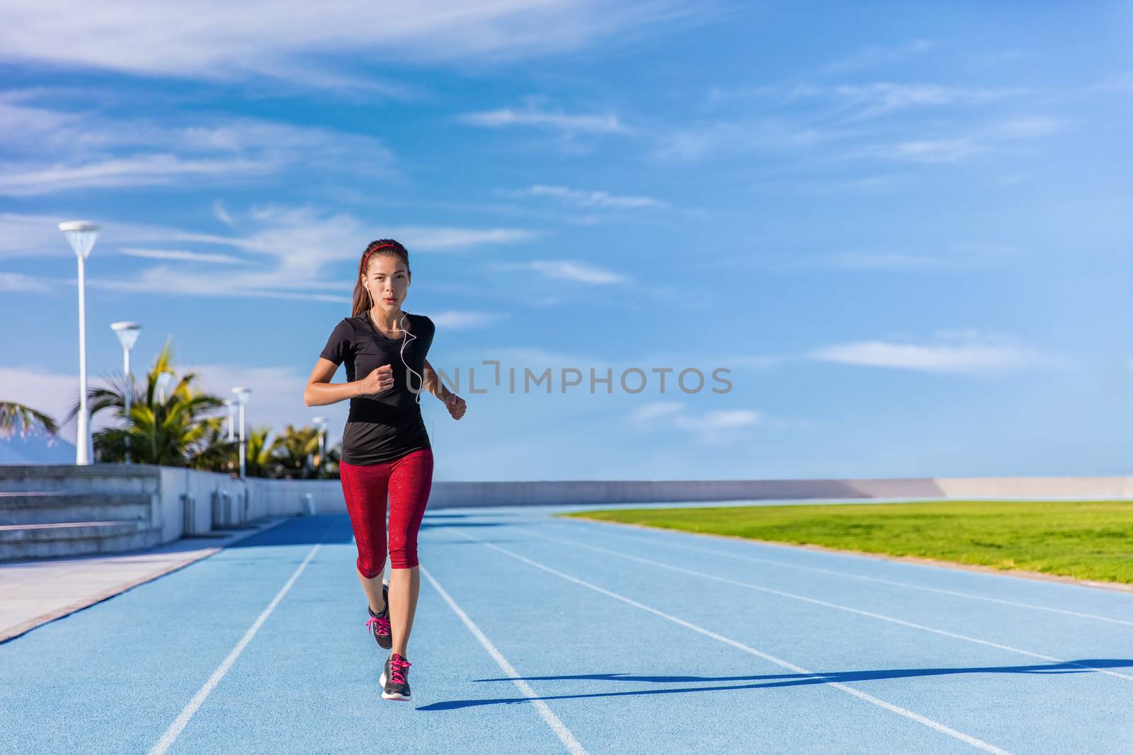 Female Asian athlete runner running on blue tracks at outdoor stadium in summer. Sporty woman jogging listening to music with earphones training cardio for weight loss success. Wellness and health.