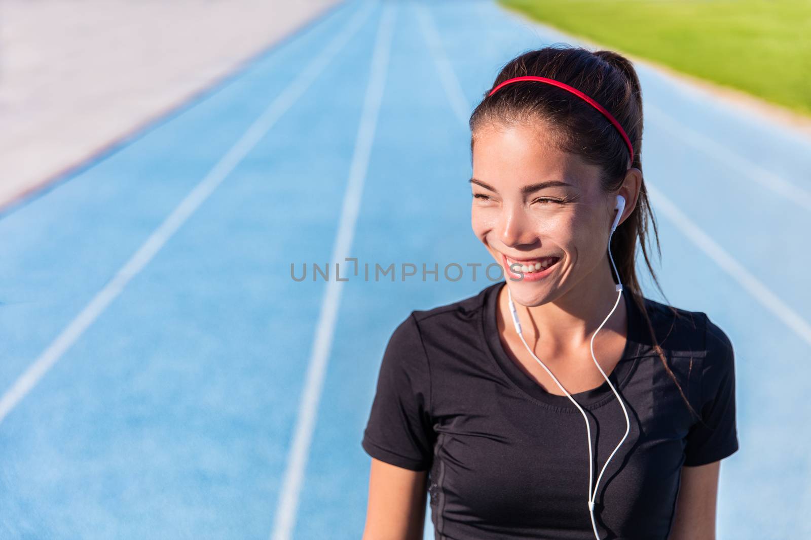 Happy track running girl runner listening to music motivation with earphones getting ready for cardio training run on blue lane athletic tracks in stadium campus. Healthy Asian athlete.