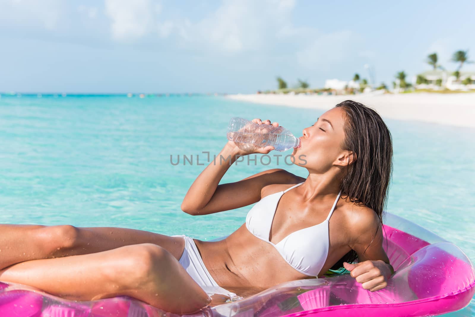Beach vacation woman on Caribbean holiday drinking fresh and clean water from plastic bottle for healthy hydration while relaxing swimming on ocean float air mattress in tropical resort.