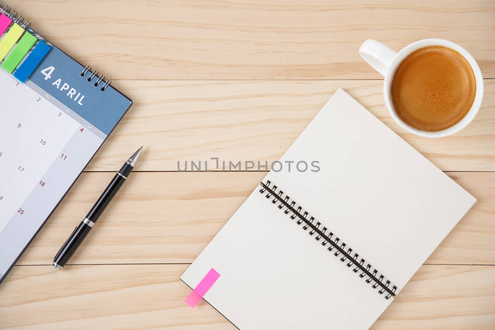 A blank notebook, a calendar, and a cup of coffee in white ceramic cup. Wooden background.