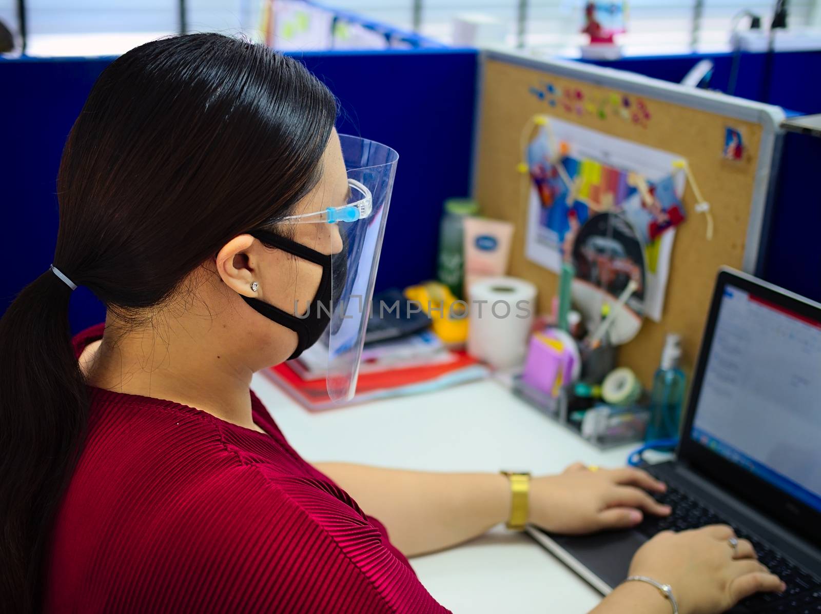 A young and beautiful Asian woman wearing a surgical mask and face shield to protect against Covid-19 while working in an office. by Faeldin