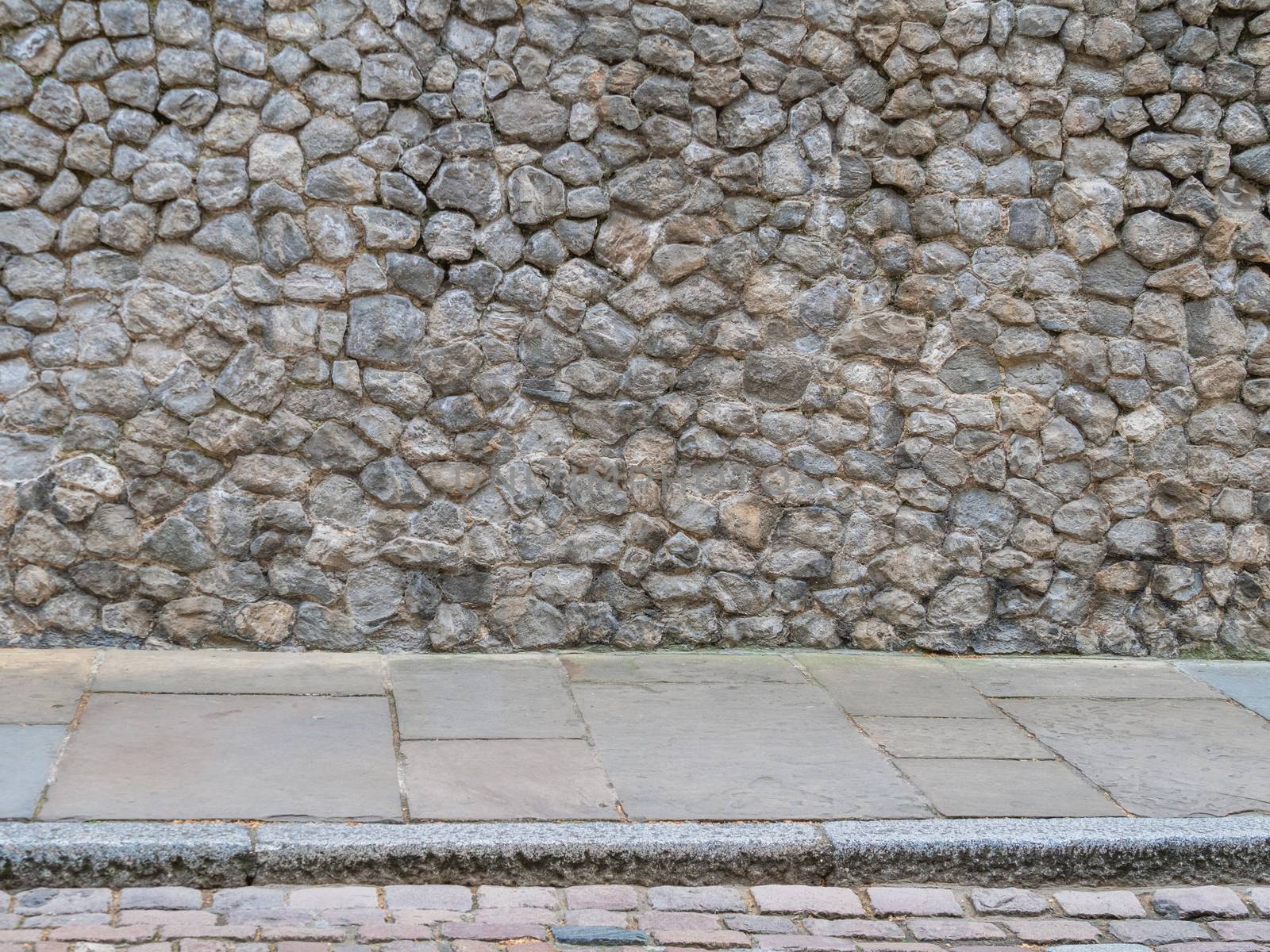 Natural rough stone wall texture background with concrete slabs and a curb on the floor. Stone wall background. Background of stone wall of boulders and pebbles. Cobble stone wall texture.
