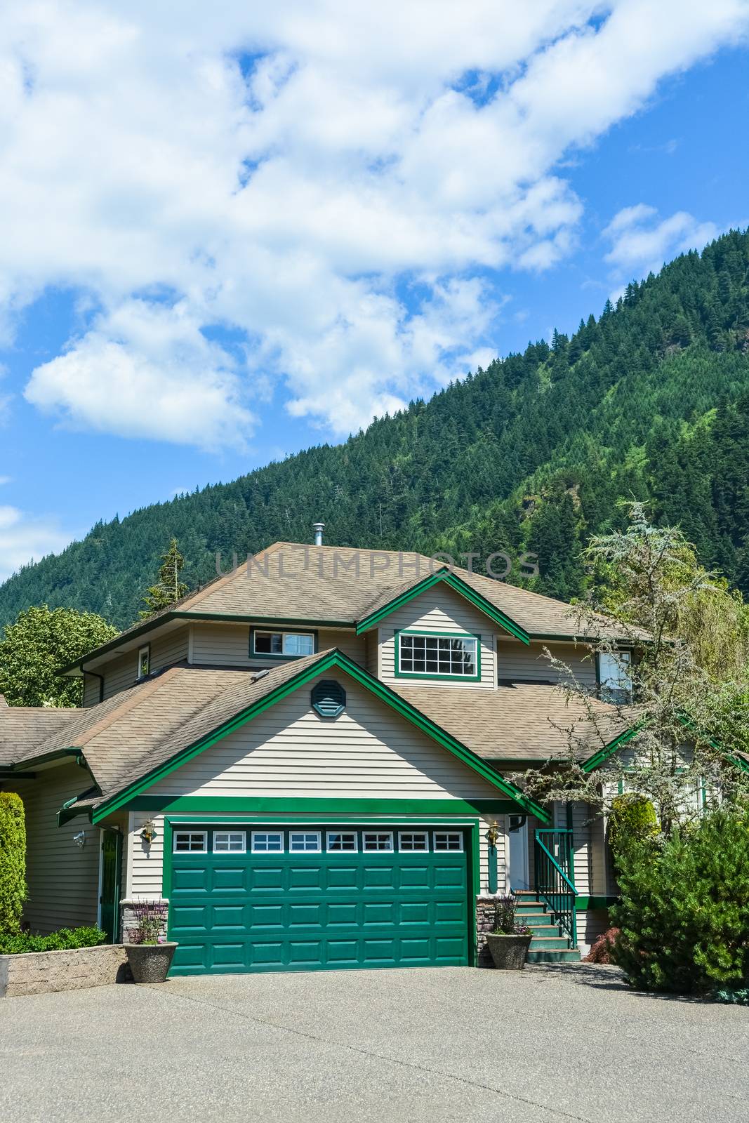 Big family house with wide concrete driveway, green wide garage door, decorated front yard. Residential house with mountain and blue sky background