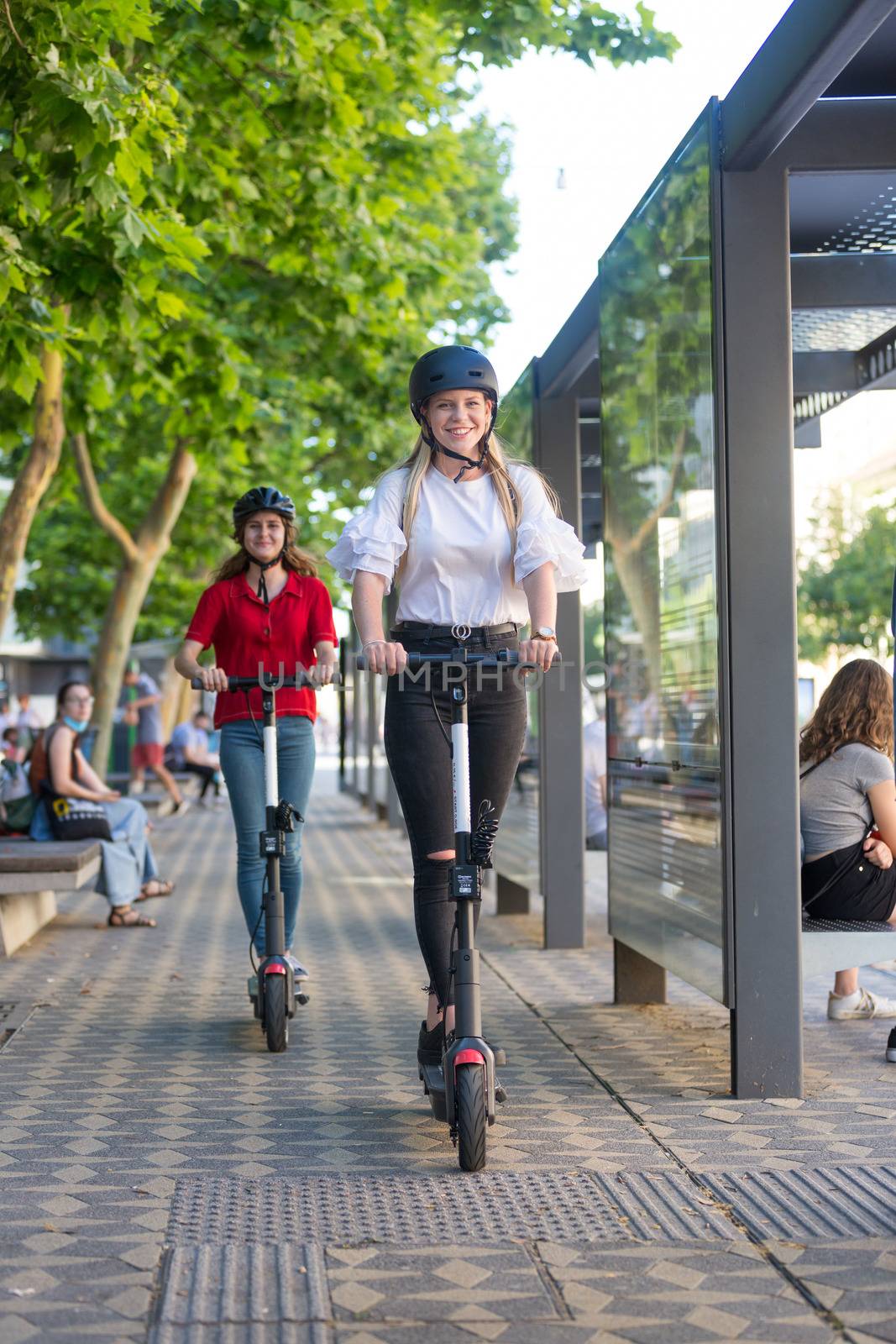 Trendy fashinable teenager girls riding public rental electric scooters in urban city environment. Eco-friendly modern public city transport by kasto