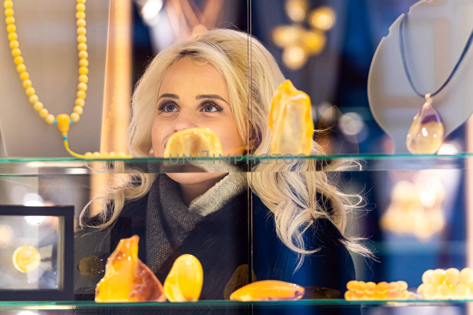 A young, beautiful, blonde woman looks at amber jewelry at a jewelry store. View through the window from the street side by askoldsb