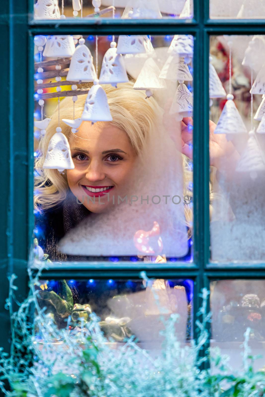 A young blonde woman looks through a window pane that is frozen and adorned with Christmas decorations. The concept of Christmas time - image