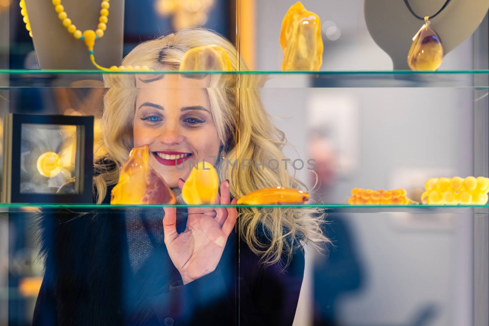 A young, beautiful, blonde woman looks at amber jewelry at a jewelry store. View through the window from the street side - image