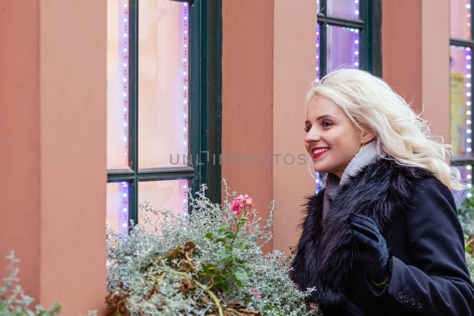 A young, smiling, blond woman from outside looks through a window adorned with Christmas decorations. The concept of Christmas time - image