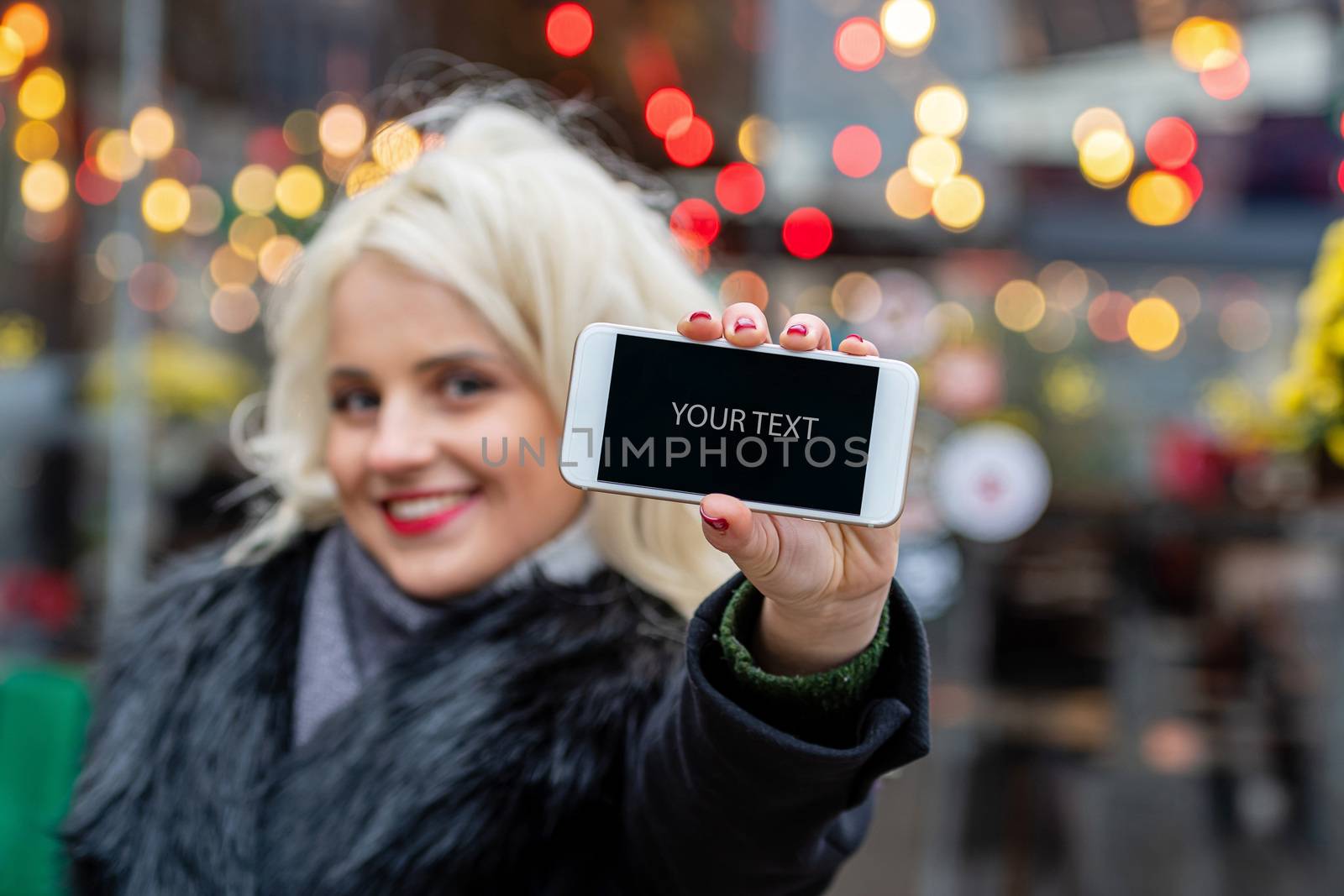 A young, beautiful blonde woman with a stretched hand shows a smartphone with a blank screen. Blurry background of holiday lights. The concept of Christmas time. Copy space - image