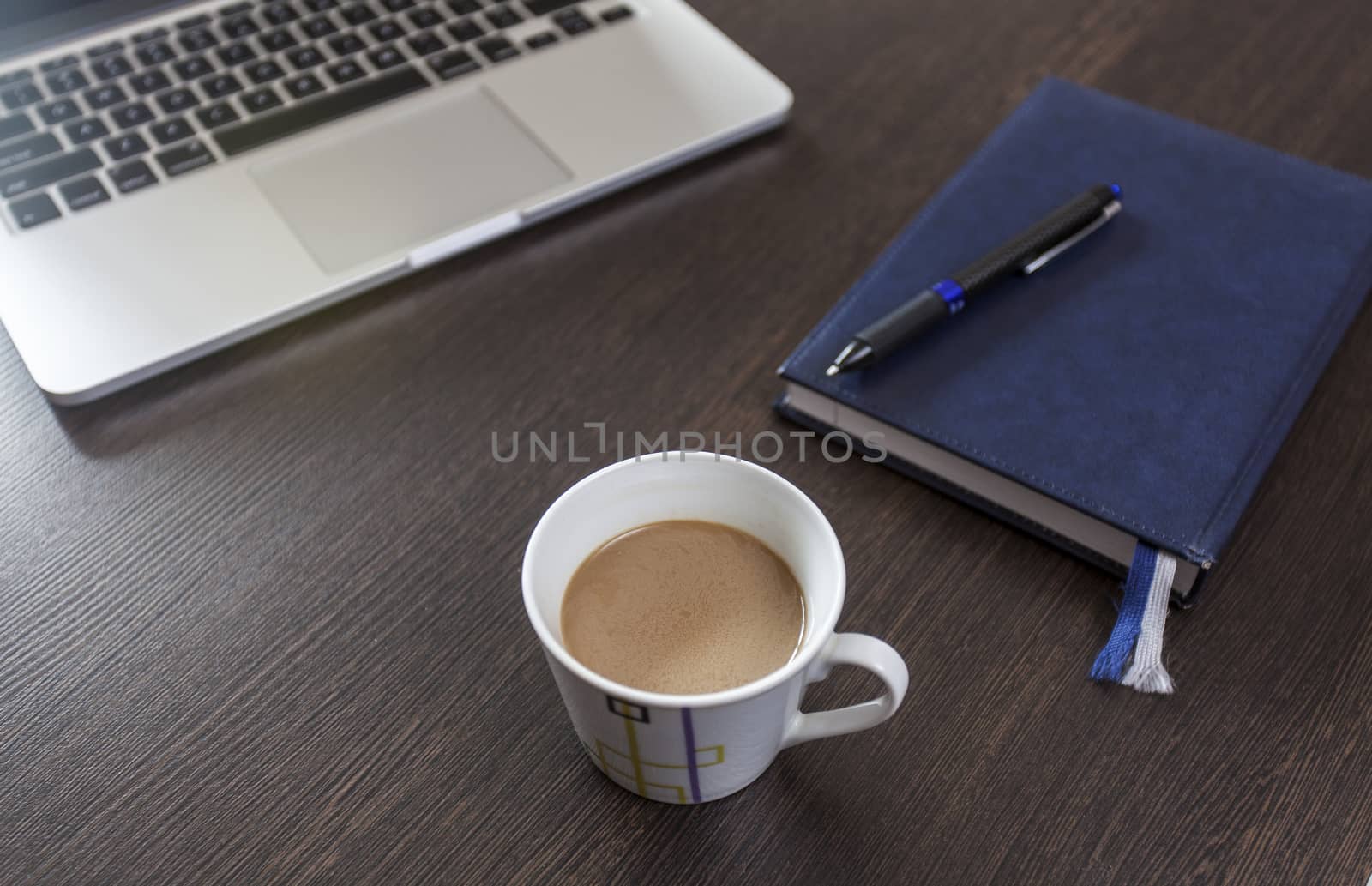 On a wooden table a laptop, a cup of coffee and a notebook