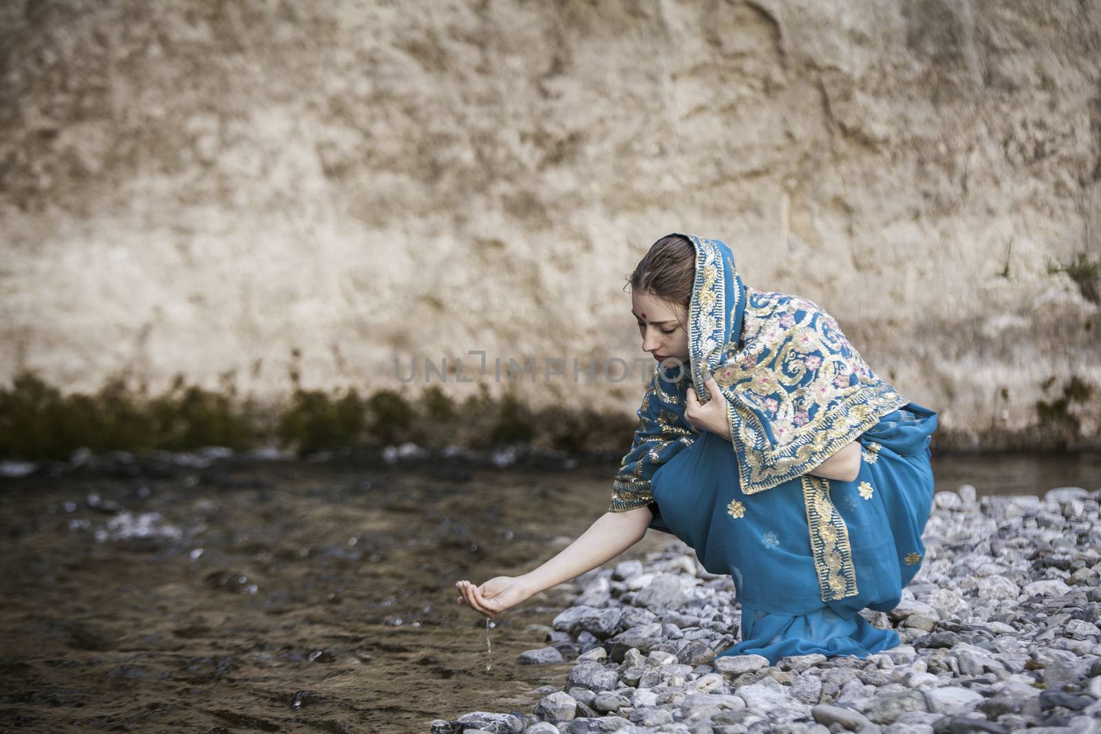 The girl in the Indian sari sits at the small river and drinks from hands