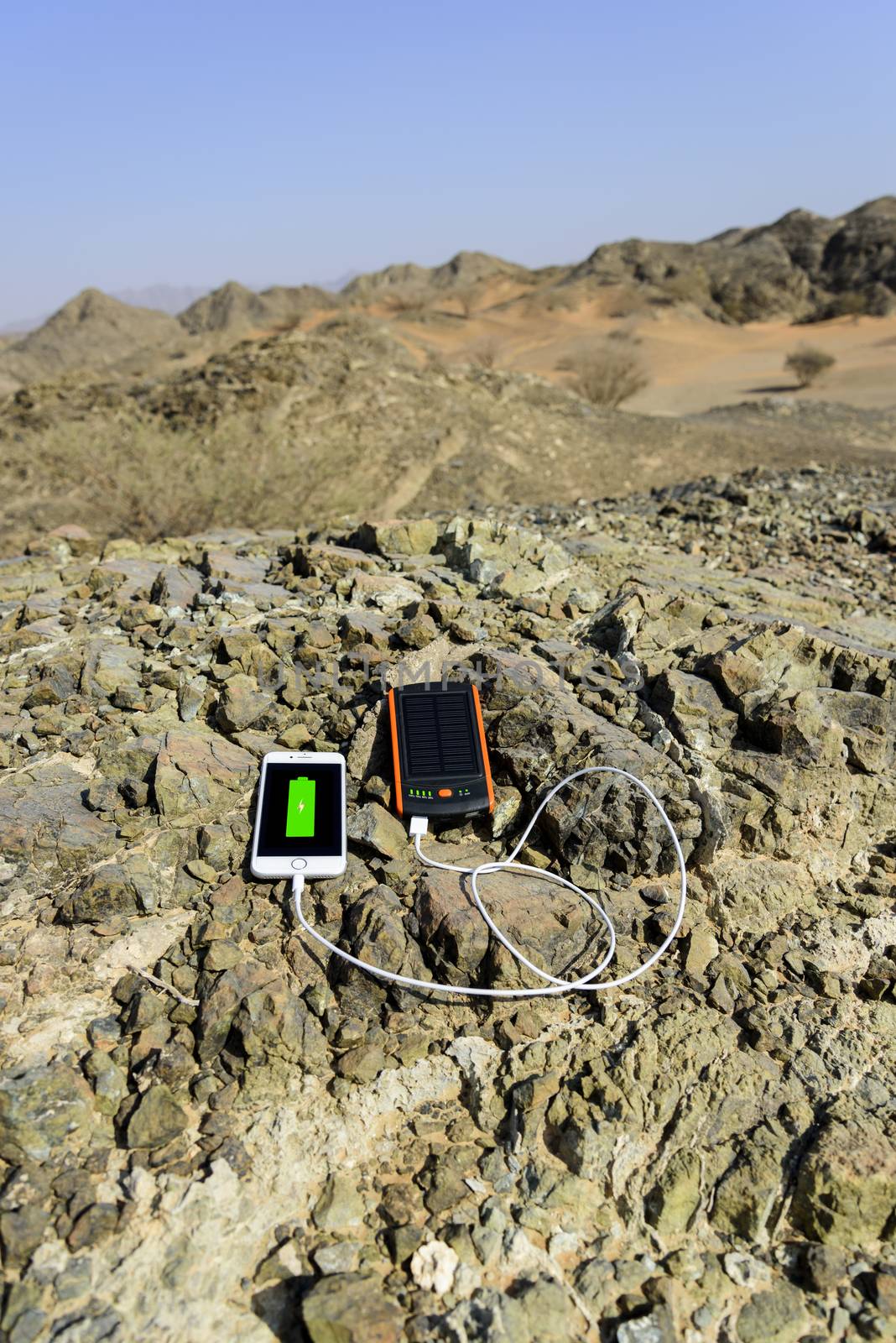 Smartphone charging with solar power bank with Dual USB port. These gadgets are lying on rocks, in the deserted mountain with some sand dunes in background.