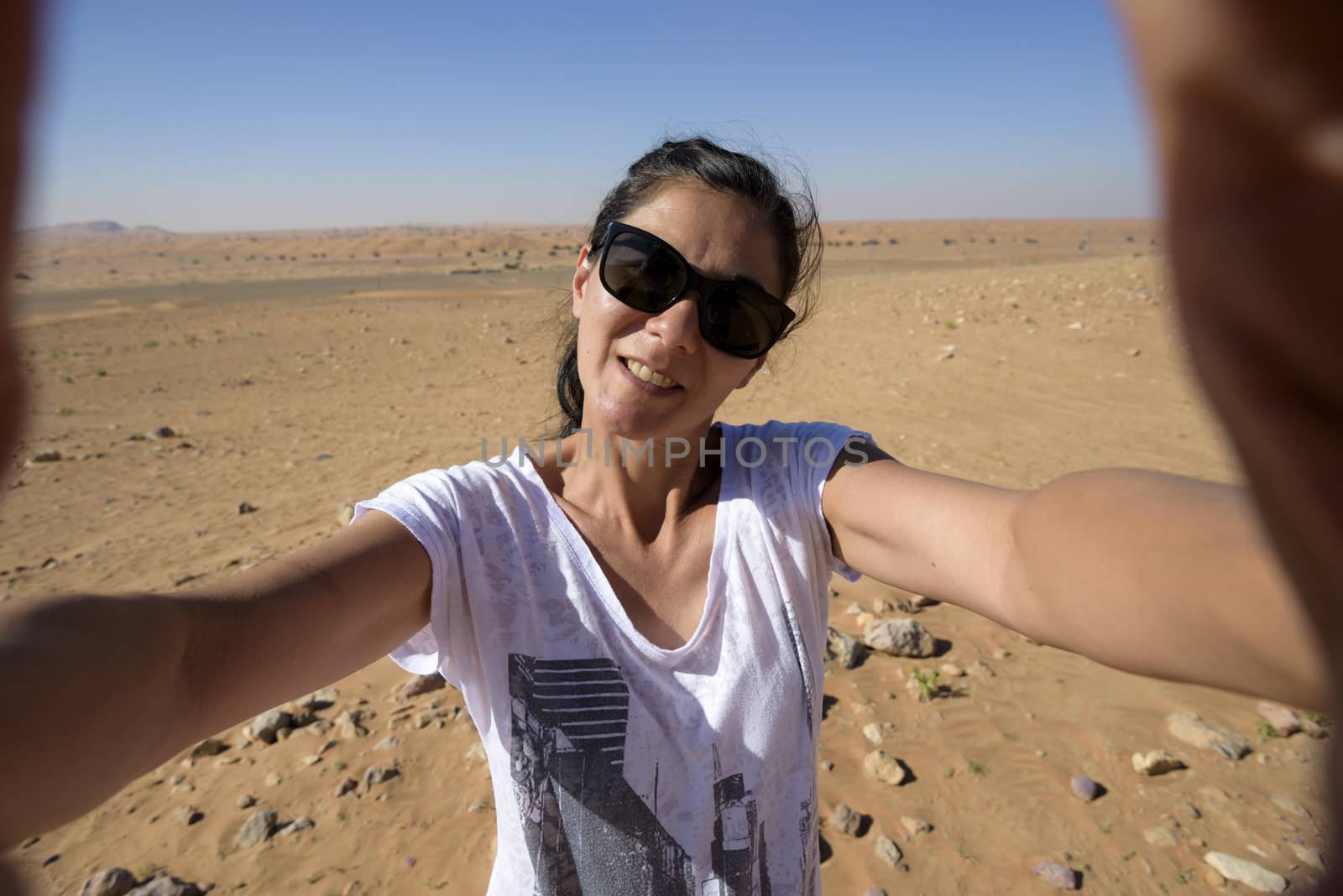 Woman taking a selfie in the desert with wide angle lens, in the 40s. We can see the desert with some dunes and stones behind her