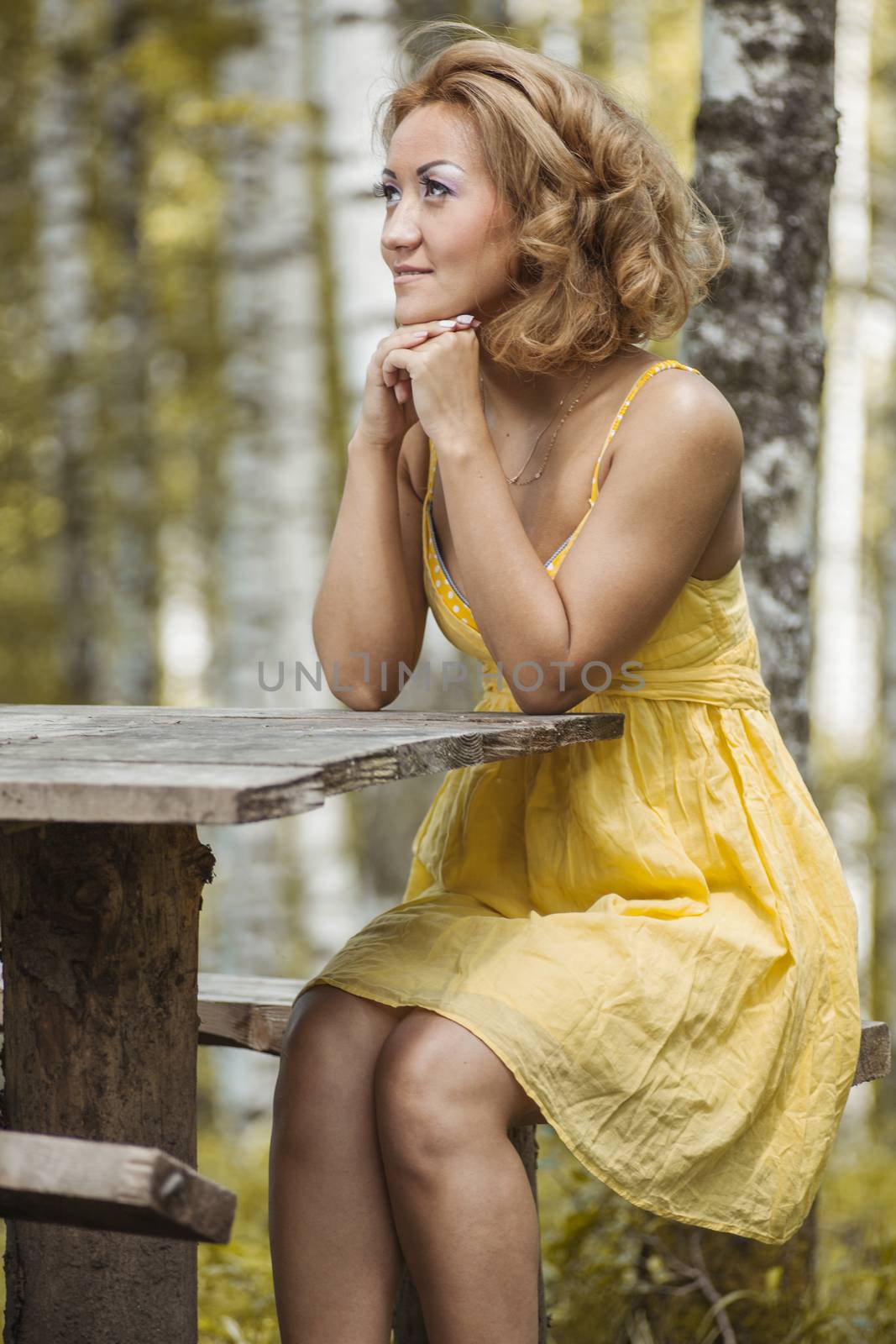 A girl in a yellow dress sitting in the woods behind a wooden table